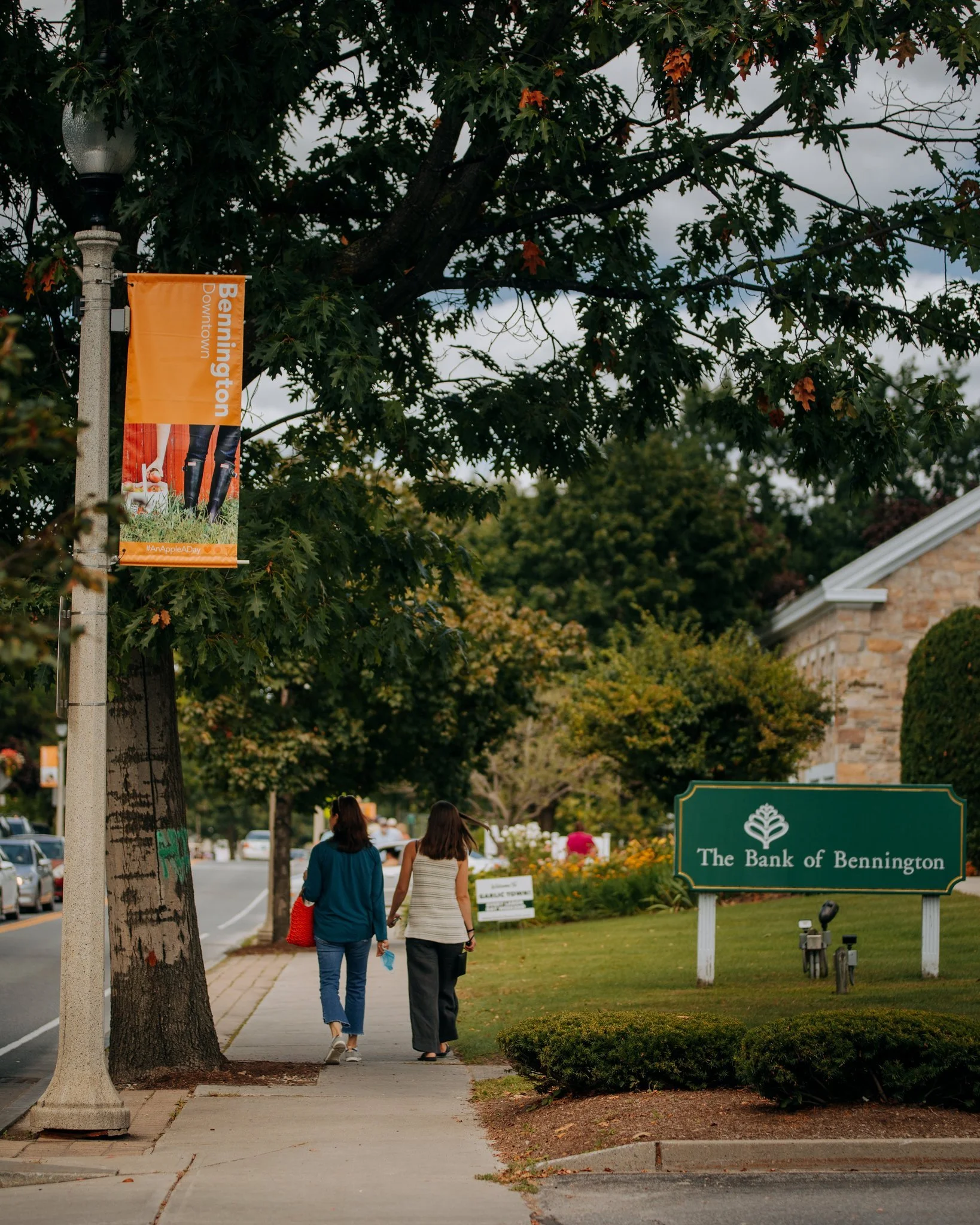 Two women walking on a sidewalk in Bennington, Vermont, near a green sign for The Bank of Bennington, with trees and parked cars along the street.