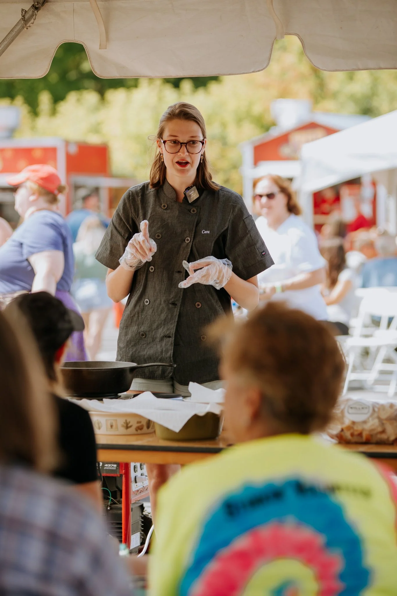 A woman wearing glasses and a dark chef's jacket, standing at a table outdoors, giving a cooking demonstration to a seated audience under a tent on a sunny day.