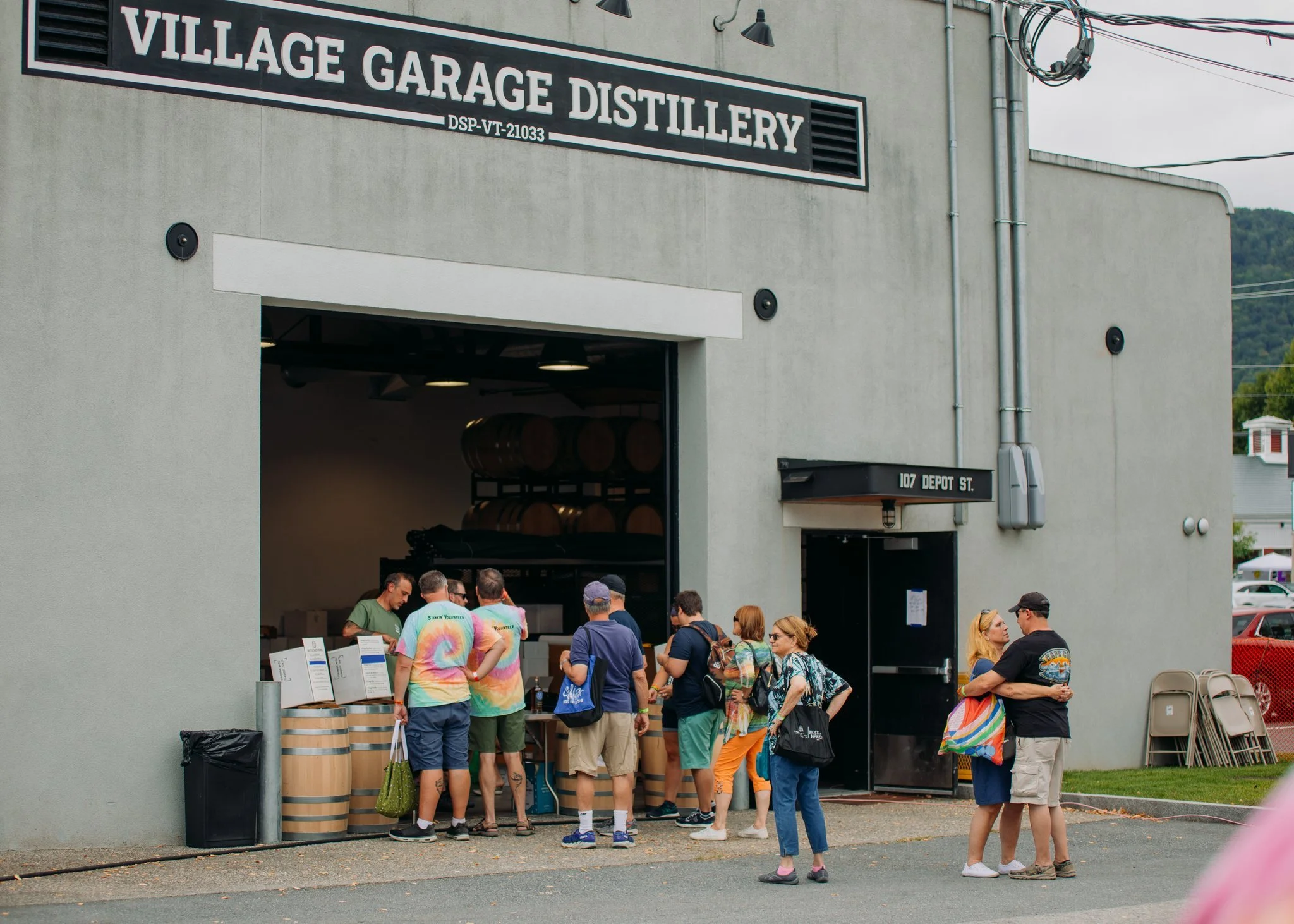 People standing outside a distillery shop with barrels visible inside. Some are waiting in line, others are talking near the entrance of Village Garage Distillery at 107 Depot Street.