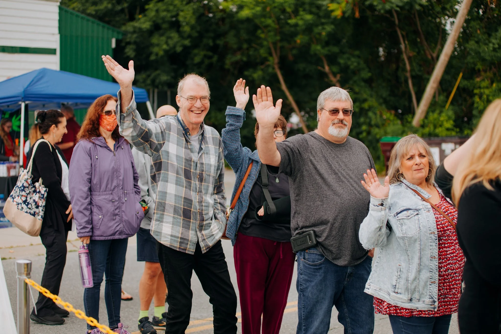 People standing in line, waving, and talking outdoors at an event, with tents and trees in the background.