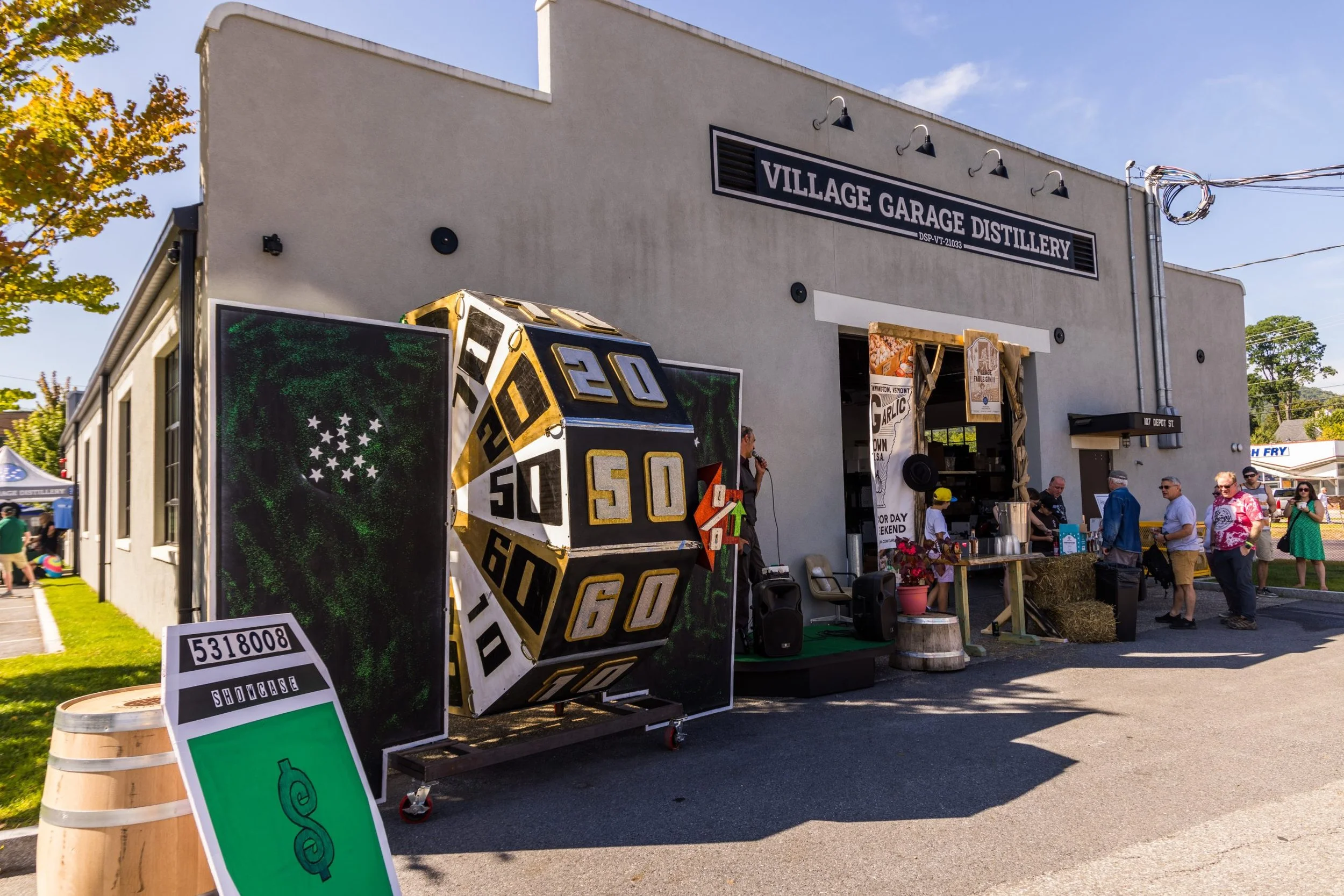 People attending an outdoor event at the Village Garage Distillery with a large colorful dice sculpture, a sign with a green dollar symbol, and a booth serving garlic-infused food. The building has a sign that reads 'Village Garage Distillery' and is