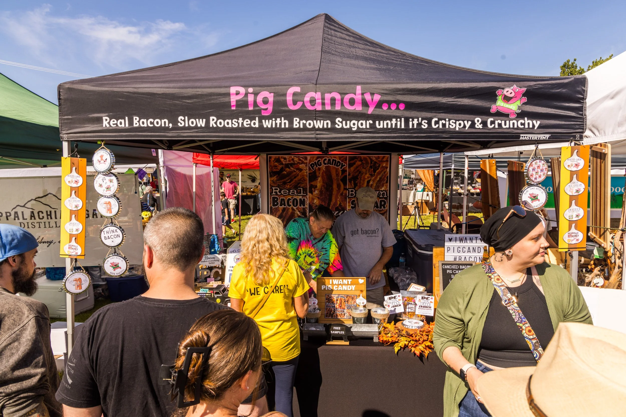 A busy outdoor market stall selling pig candy, with people gathered around. The stall has a sign that reads 'Pig Candy... Real Bacon, Slow Roasted with Brown Sugar until it's Crispy & Crunchy' and features a cartoon pig character. Several decorative 