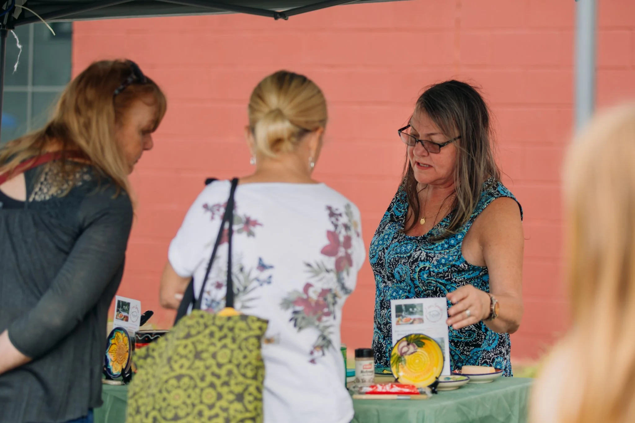 A woman with glasses and a blue patterned top demonstrating or explaining something at a table with colorful plates and small jars, while three women listen attentively at an outdoor event against a peach-colored wall.