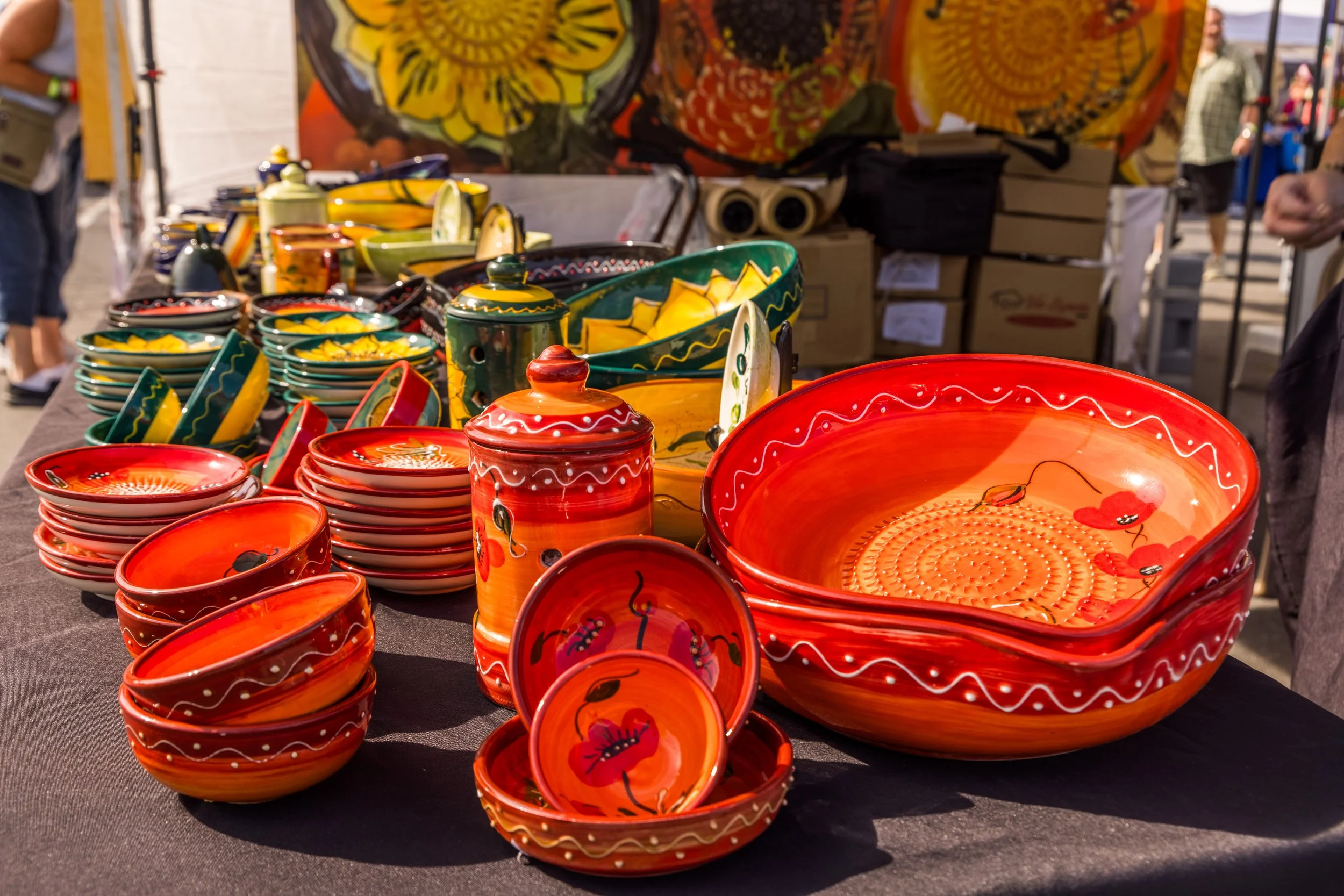 Colorful handcrafted pottery including bowls, plates, and a large dish with floral and abstract patterns on display at an outdoor market.