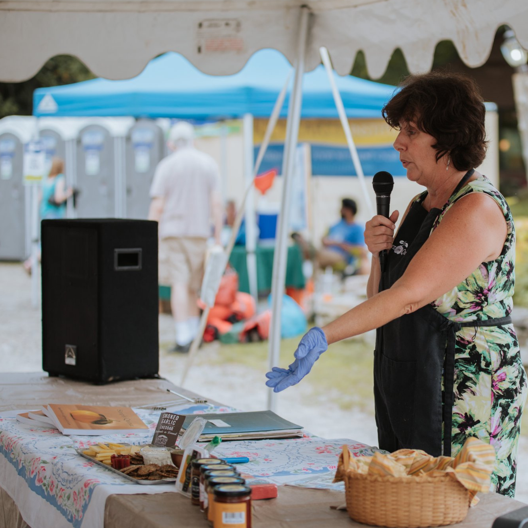 A woman in a floral dress and apron speaking into a microphone at an outdoor event. She is standing under a white tent next to a table covered with a tablecloth, food, and condiments. She wears a blue glove on one hand.