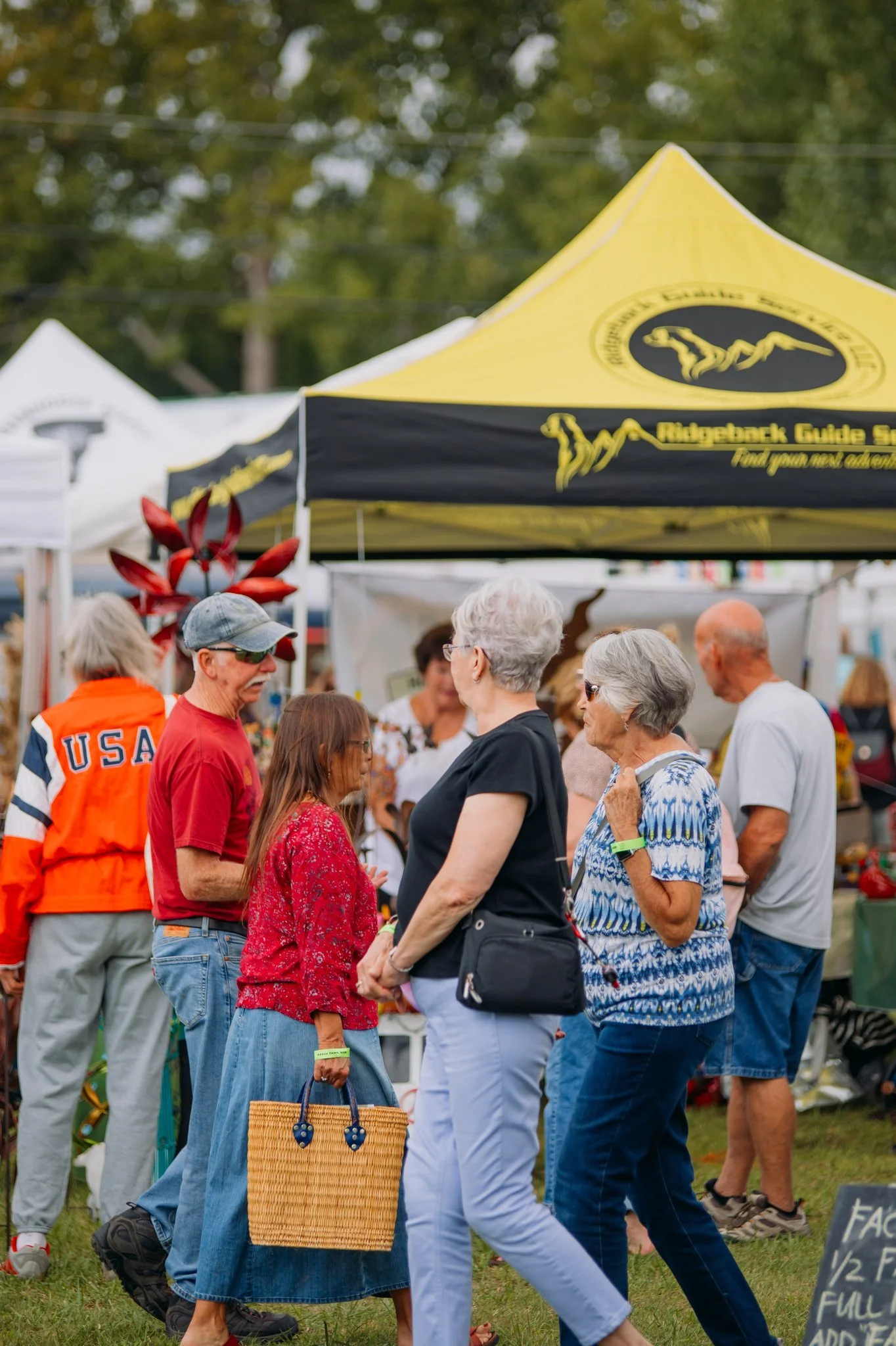 People gathering at an outdoor market or festival with vendor tents, including yellow and white ones. The scene includes older adults, a woman with a woven bag, and a person wearing a jacket with 'USA' on it.