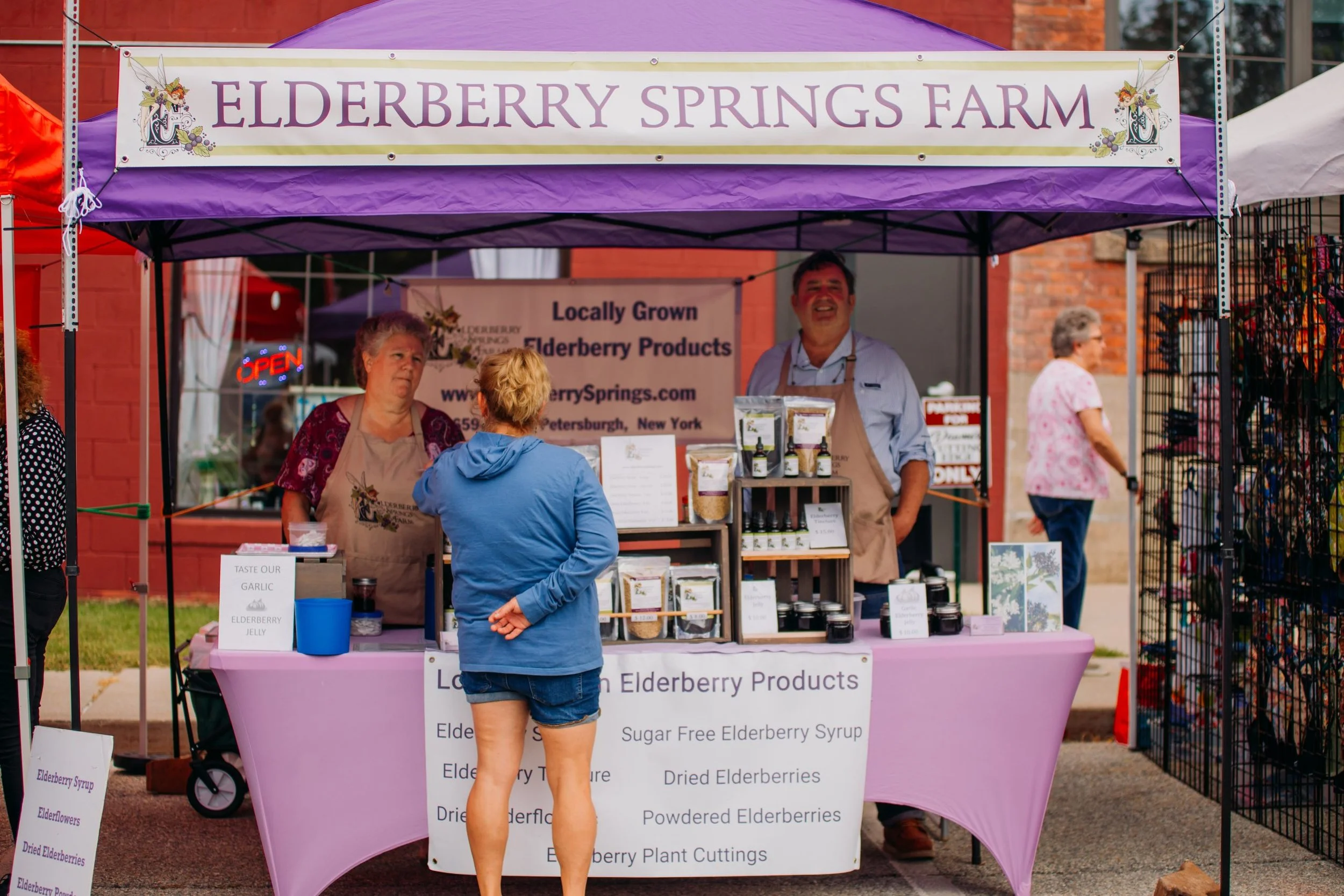 A woman and a man are behind a booth selling elderberry products at an outdoor market. The booth has a purple canopy with a sign reading 'Elderberry Springs Farm.' The woman is wearing an apron, and the man is smiling and wearing an apron over a ligh