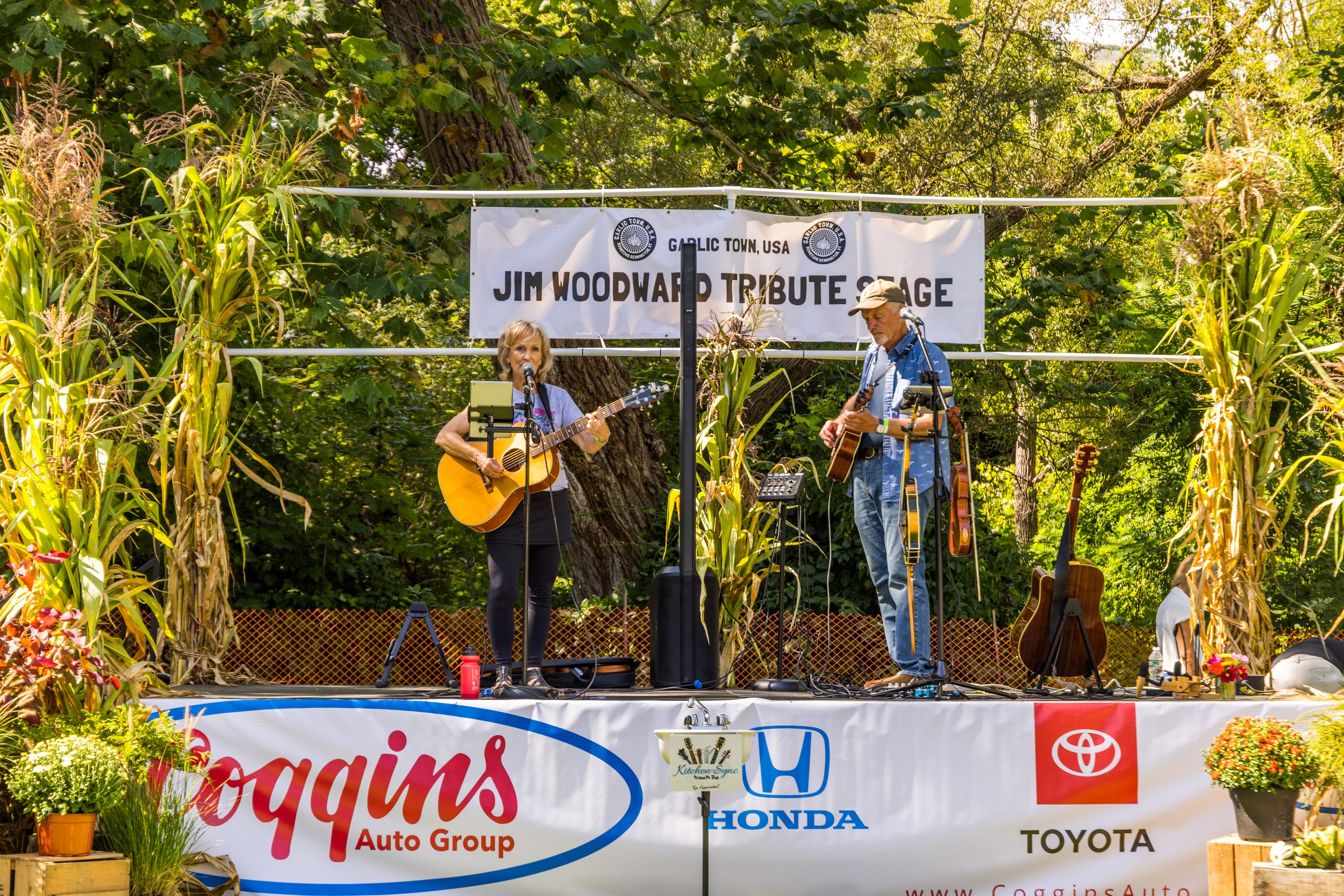 Two musicians playing guitars on an outdoor stage decorated with cornstalks and surrounded by trees. The woman on the left is singing into a microphone, and the man on the right is focused on playing his guitar. A banner above reads 'Jim Woodward Tri