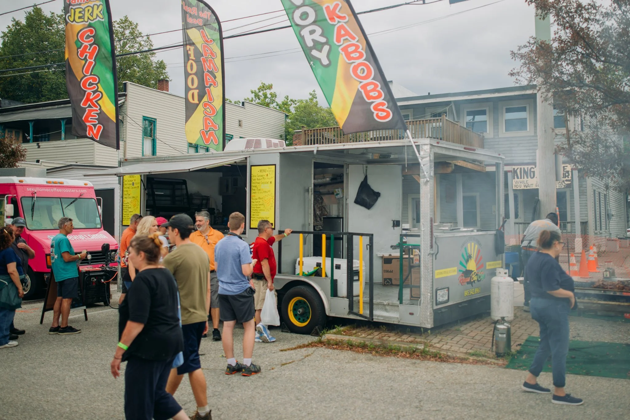 A food truck selling baked goods and fried food at an outdoor event. There are colorful flags advertising chicken, jalapeno, and kabobs. People are standing in line and chatting, with some ordering food from the truck.