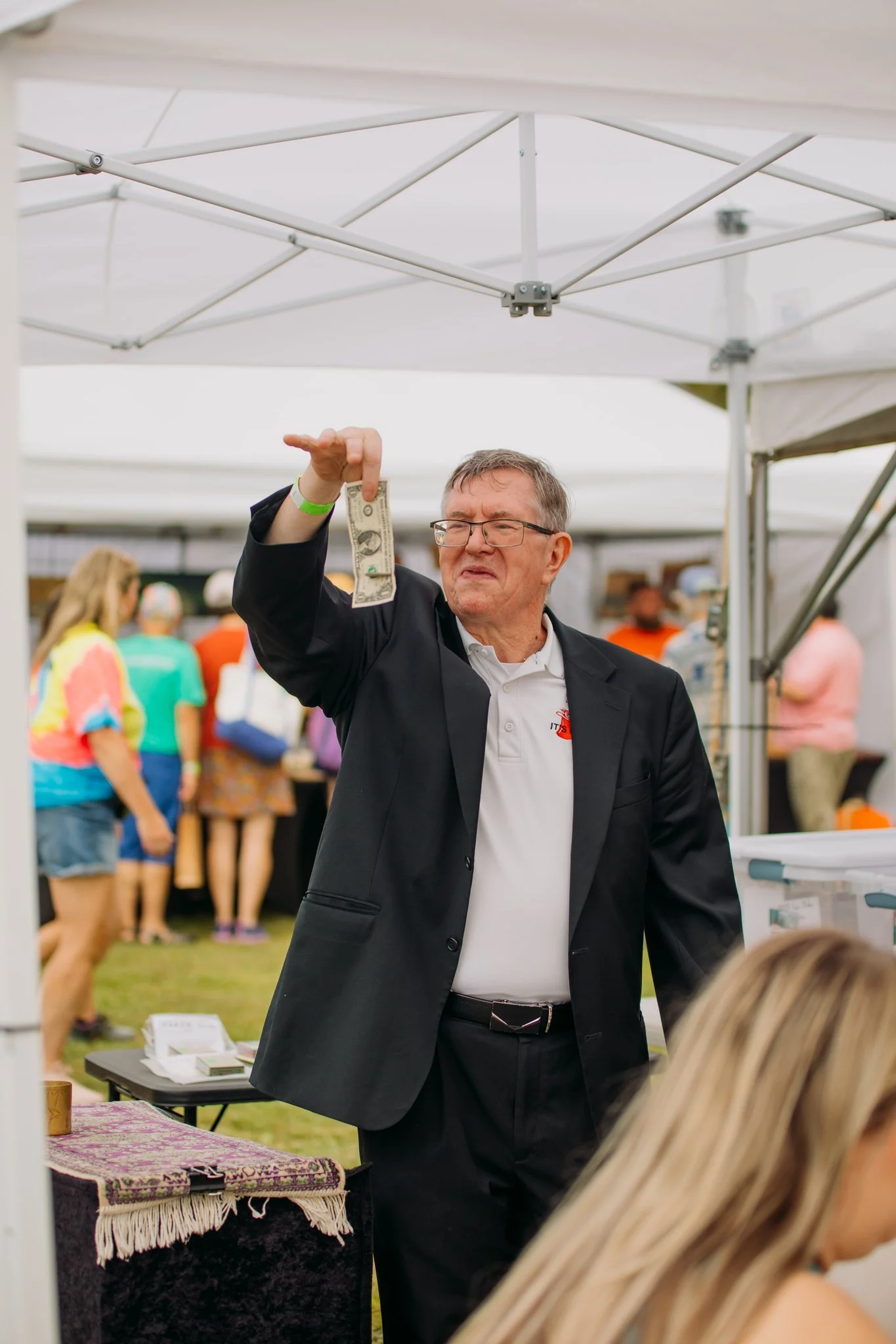An older man in a black blazer and glasses holding a dollar bill, making a displeased face at an outdoor event with white tents and people in colorful clothing in the background.