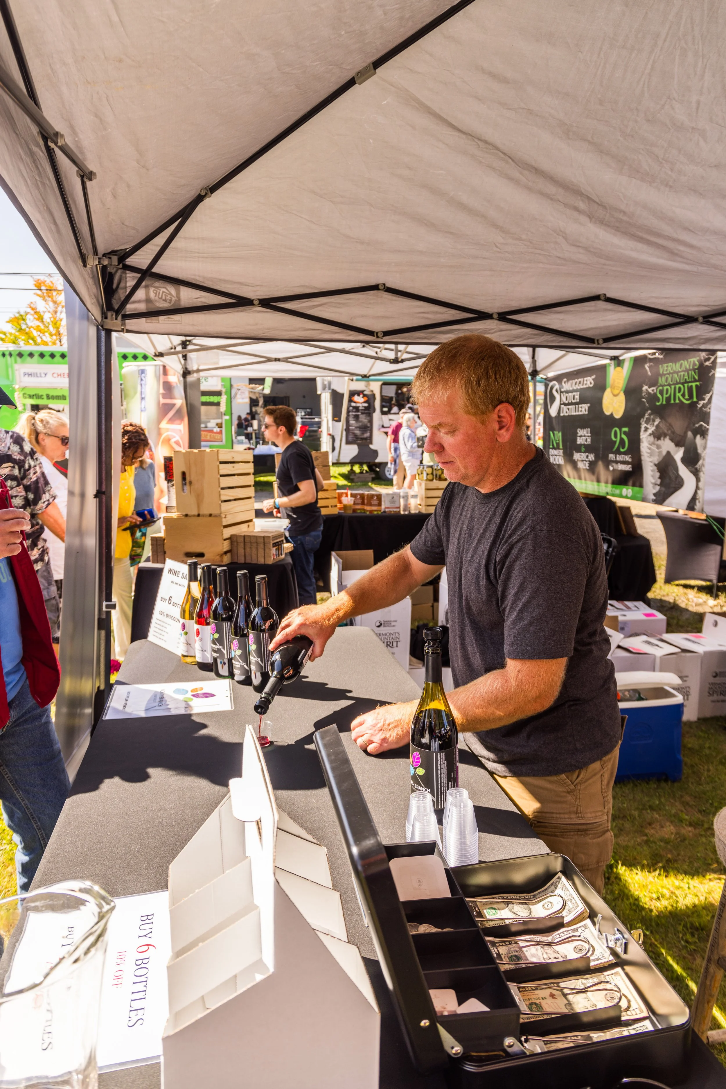 A man pouring wine at an outdoor market booth with bottles of wine on display and a sign that says 'Buy 6 Bottles'.