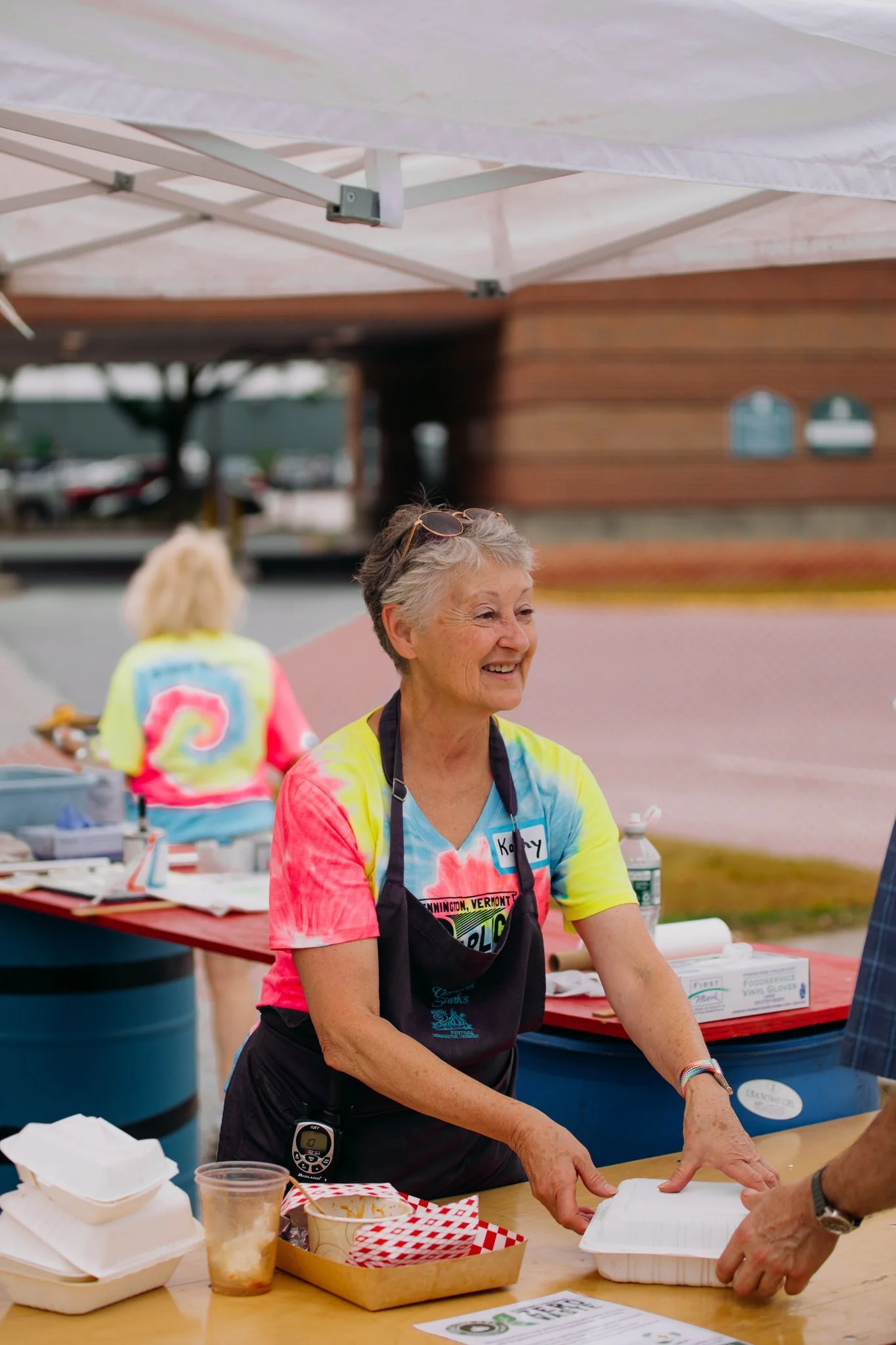 A smiling older woman wearing a colorful tie-dye t-shirt and black apron, standing at a table outdoors, handing a food container to someone, with a tie-dye shirted woman in the background and a tent overhead.