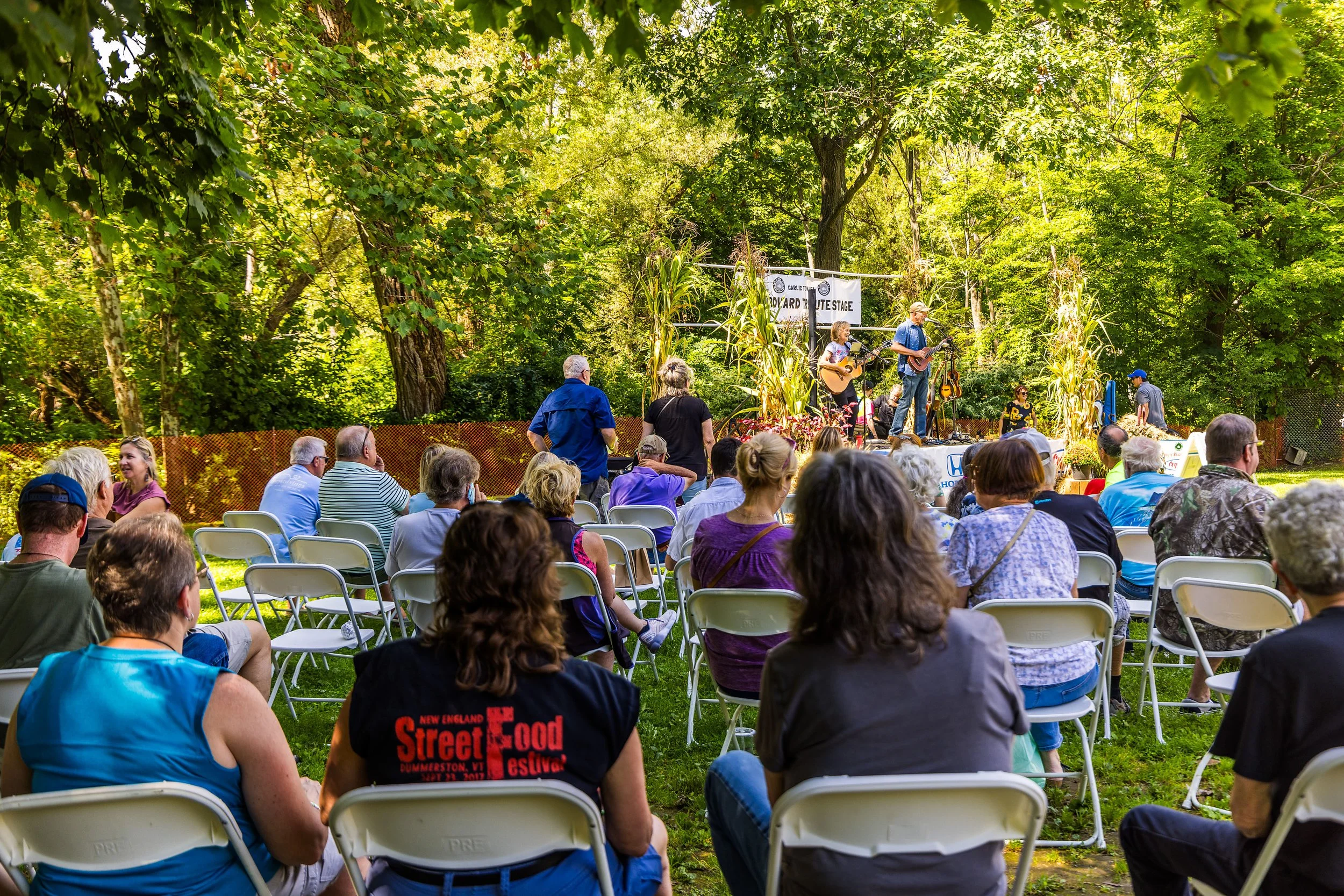 Outdoor music concert with audience seated on white chairs watching performers on stage surrounded by trees and greenery.