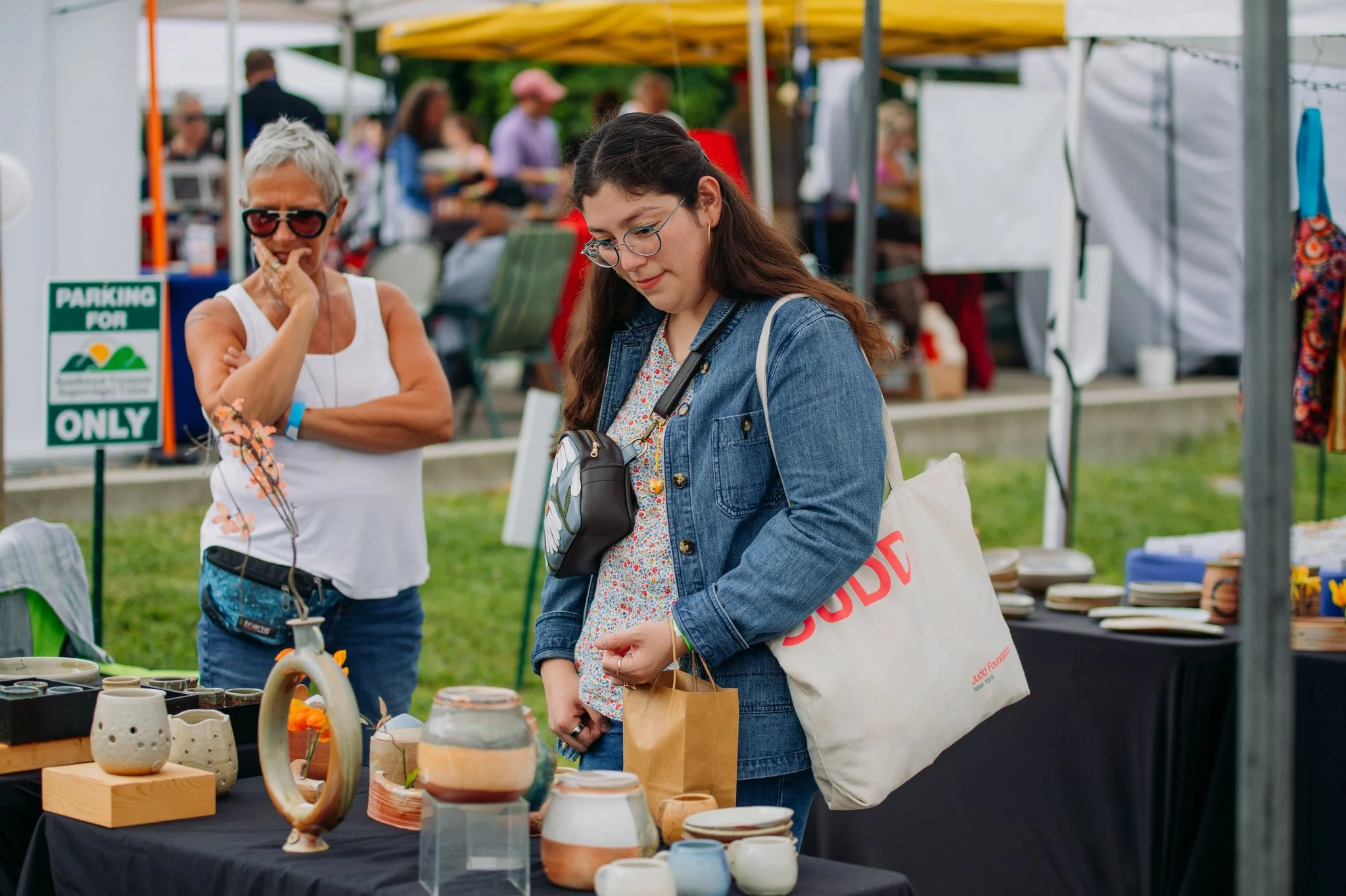 A young woman with glasses and long brown hair shopping at an outdoor craft fair, looking at pottery on a table. An older woman with short gray hair, wearing sunglasses, a white tank top, and a blue fanny pack, stands nearby with her hand on her chin