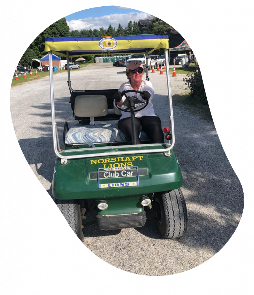 A woman sitting in a green golf cart with a yellow and blue roof, holding the steering wheel. The cart has several signs on the front, including "Norshaft Lions," "Club Car," and Lions Club logos. She is outdoors on a gravel path with trees, buildings, and traffic cones in the background.