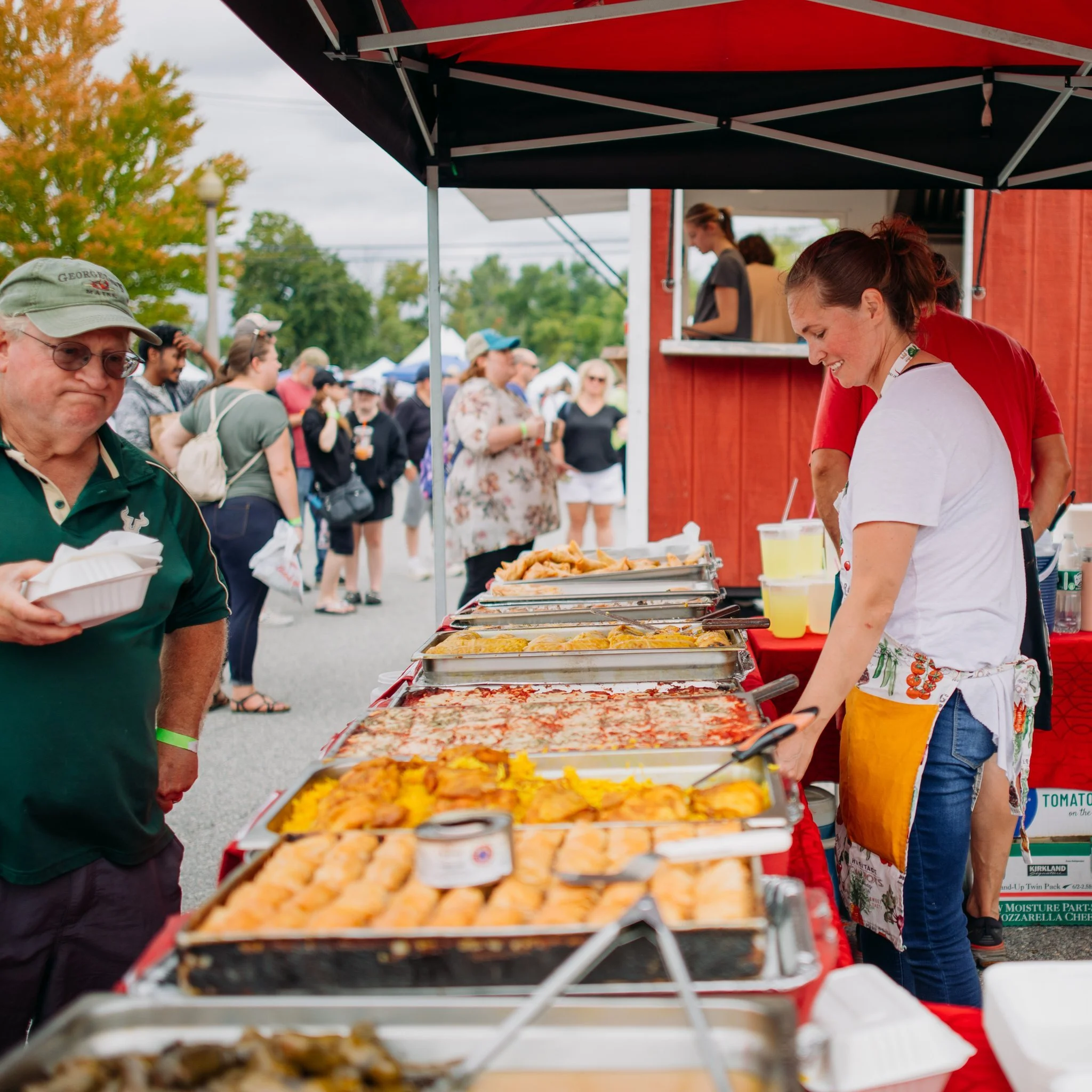 People at a food stand with trays of buffet-style dishes at an outdoor event, surrounded by festival attendees.