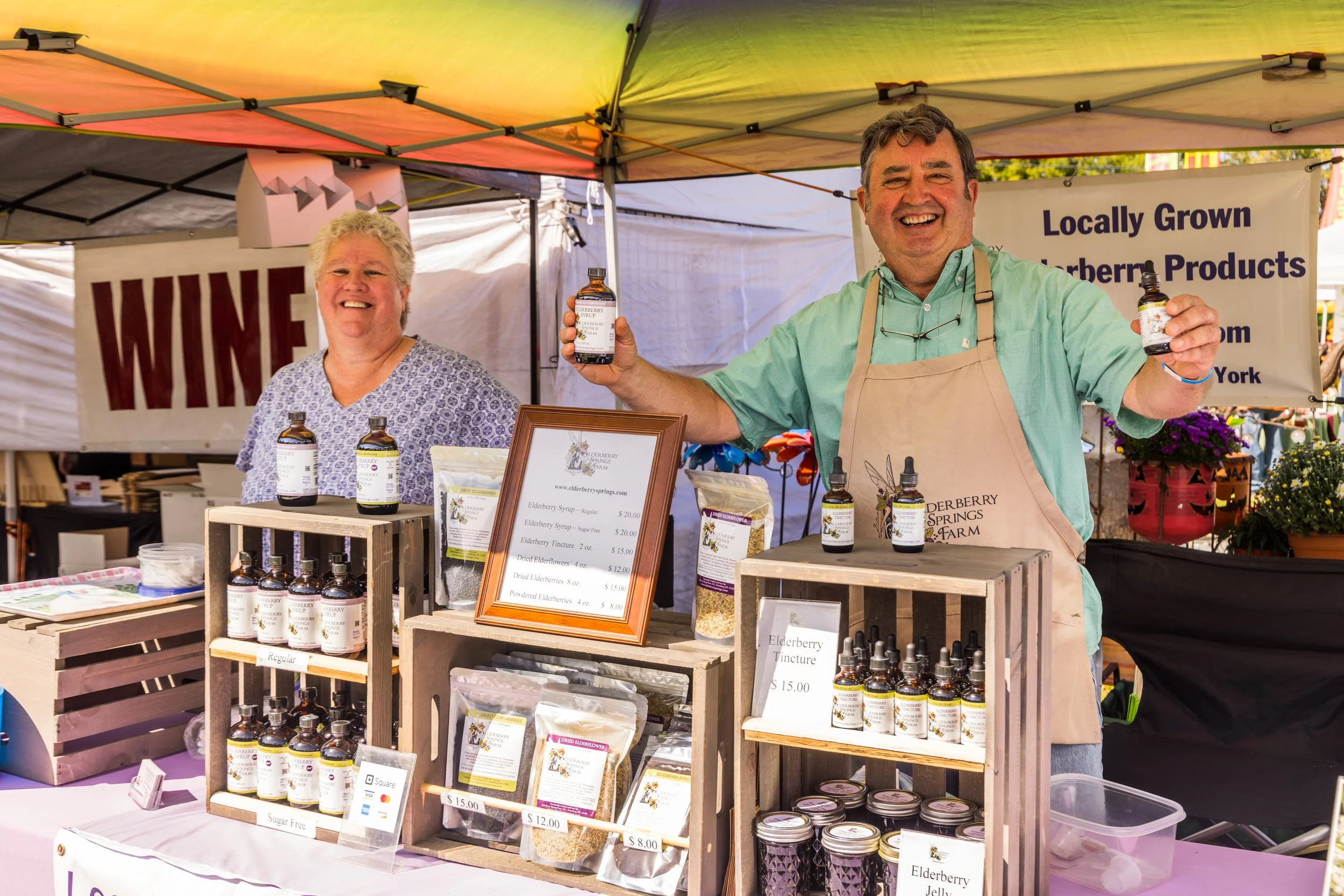 A man and woman at a booth selling elderberry products, with the man holding elderberry syrup bottles and smiling, under a yellow and green canopy with signs for wine and elderberry products.