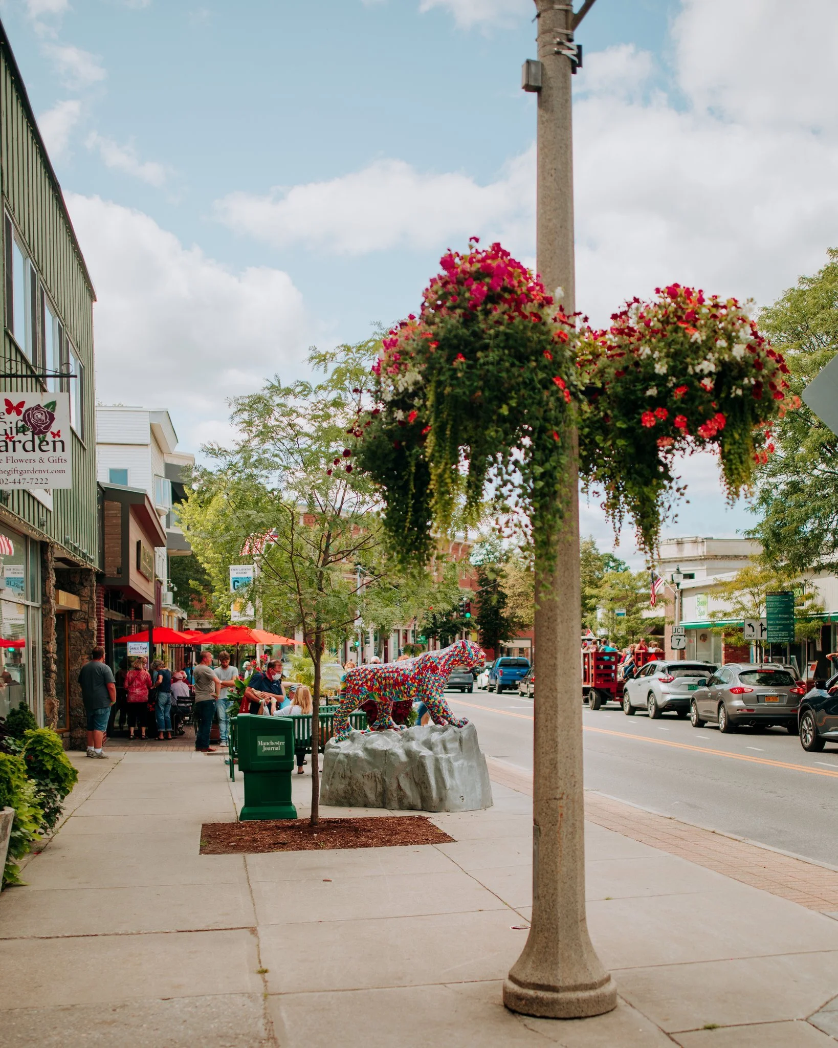 A city sidewalk with people dining outside at a restaurant, a colorful flower sculpture of a lion, a hanging flower basket on a streetlight, and parked cars along the street on a partly cloudy day.