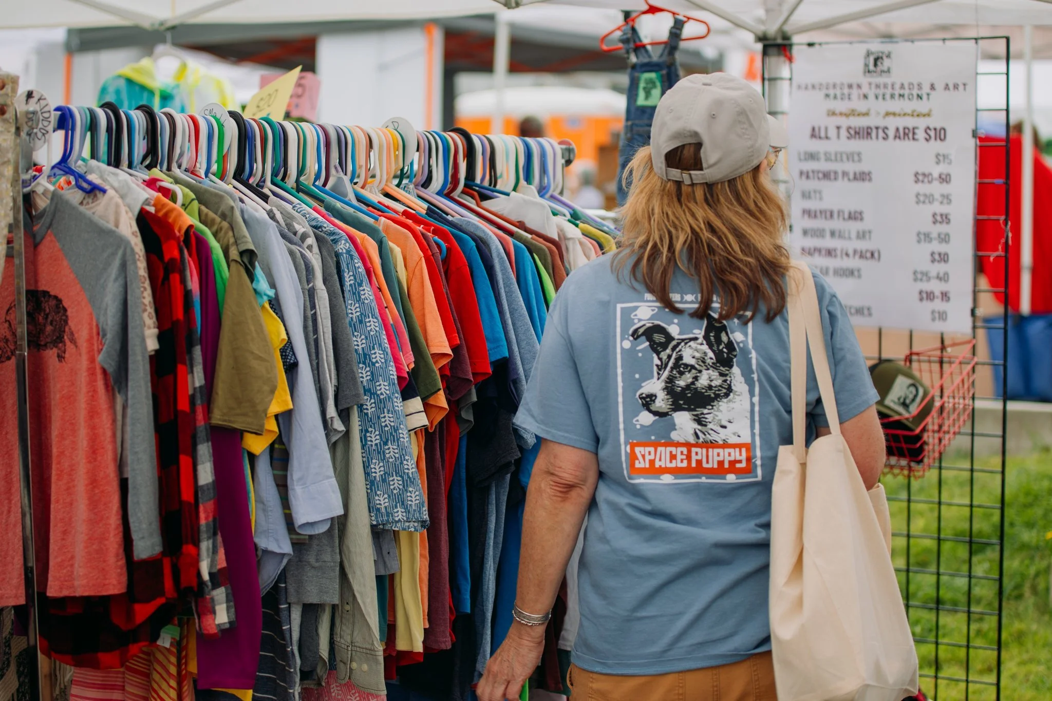 A woman with red hair wearing a gray cap, a light blue T-shirt with a dog graphic and the words 'Space Puppy,' is browsing through a rack of colorful T-shirts at an outdoor market. There is a sign listing prices of various items behind her.