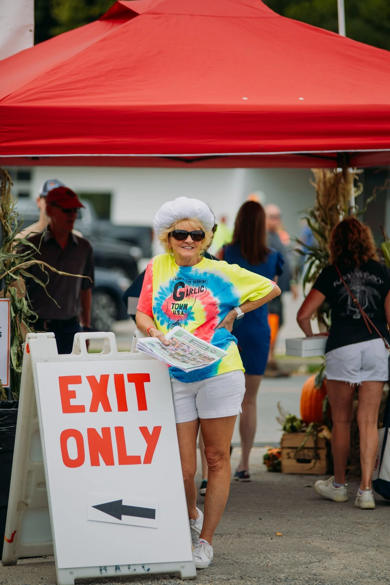 A woman with white hair, sunglasses, and a tie-dye T-shirt, smiling and holding newspapers, standing near an 'EXIT ONLY' sign at an outdoor event or market under a red canopy. There are other people and pumpkins in the background.