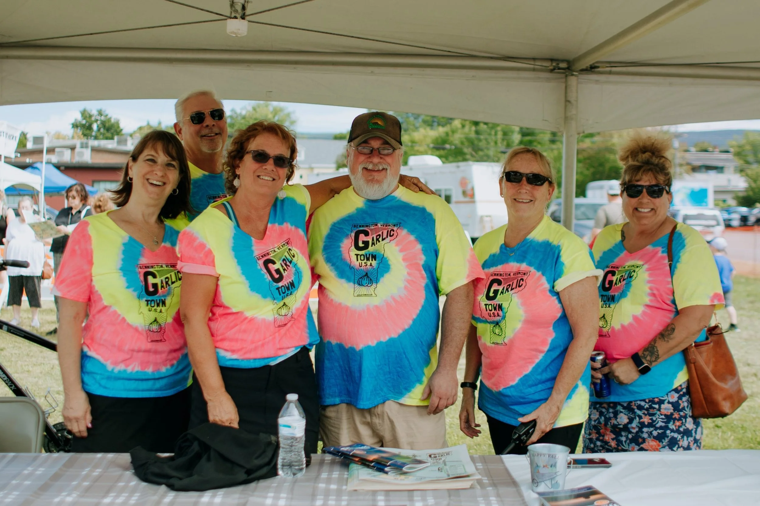 Group of six people wearing matching colorful tie-dye shirts with the text 'Garlic Town U.S.A.' smiling and standing under a canopy at an outdoor event.