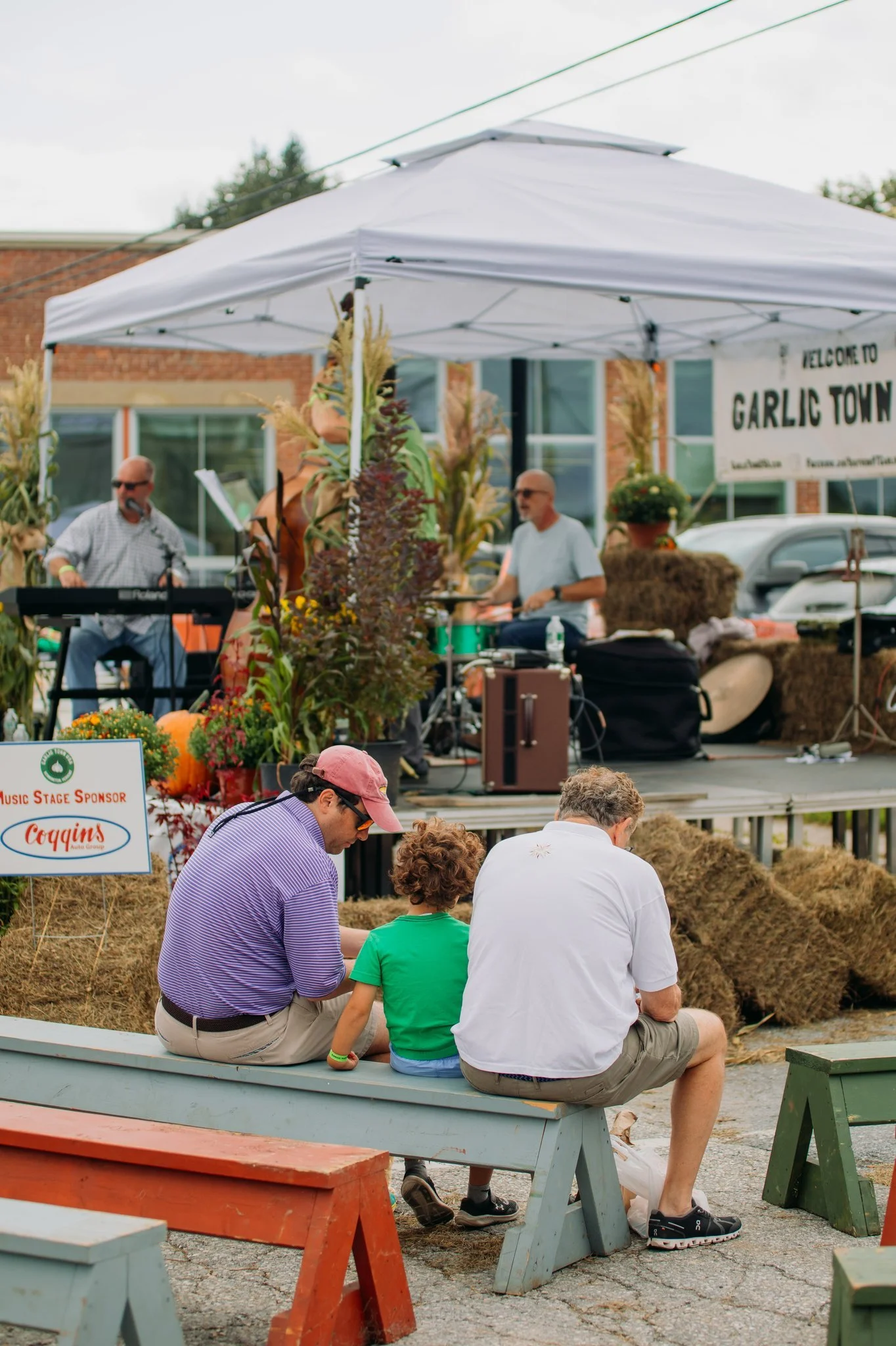 A small outdoor music event titled 'Garlic Town,' with a band performing on a stage decorated with corn stalks and hay bales, and people sitting on benches in front.