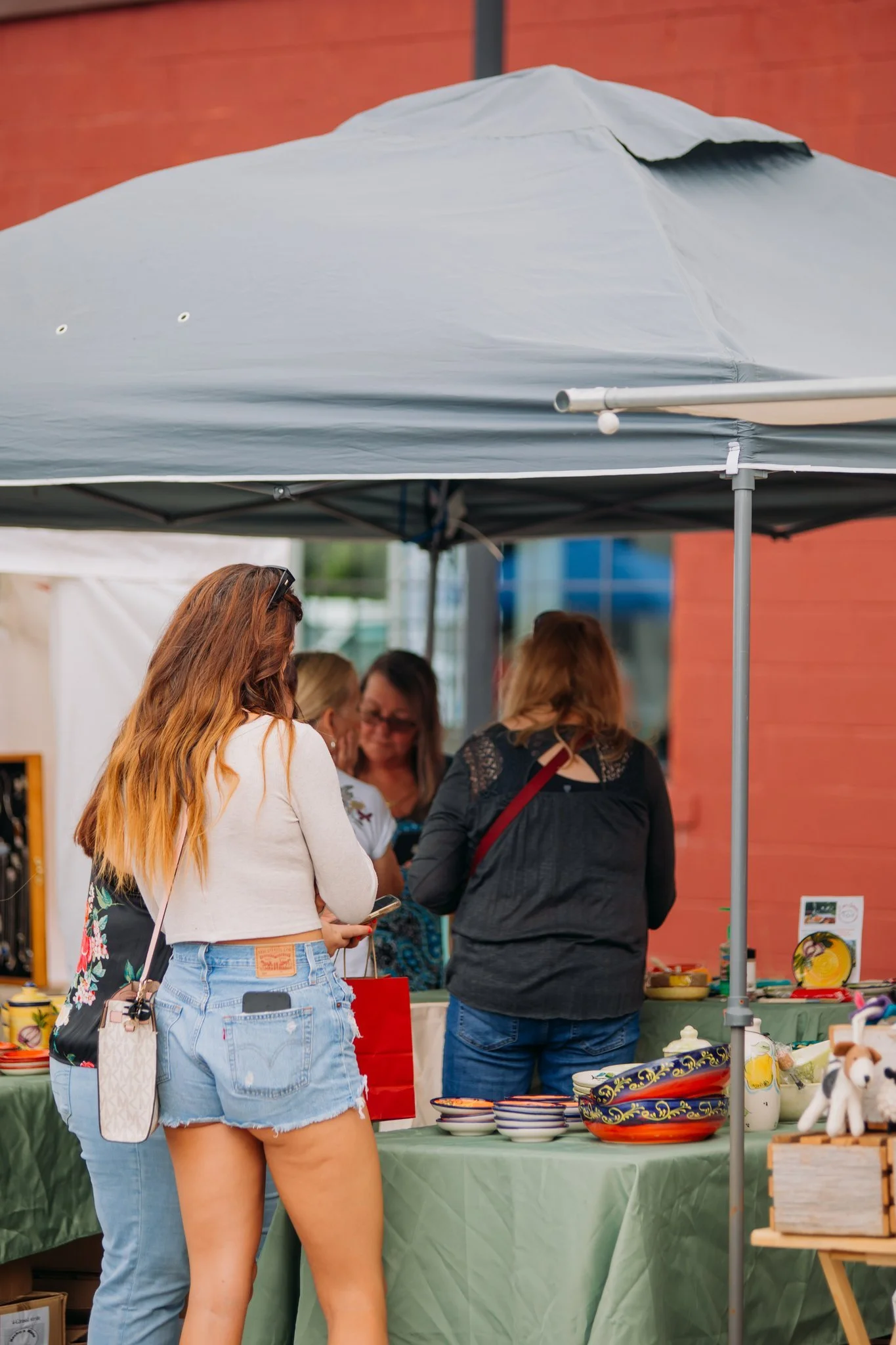 People shopping at an outdoor market stall under a large gray canopy tent, with various ceramic bowls and decorative items on display.