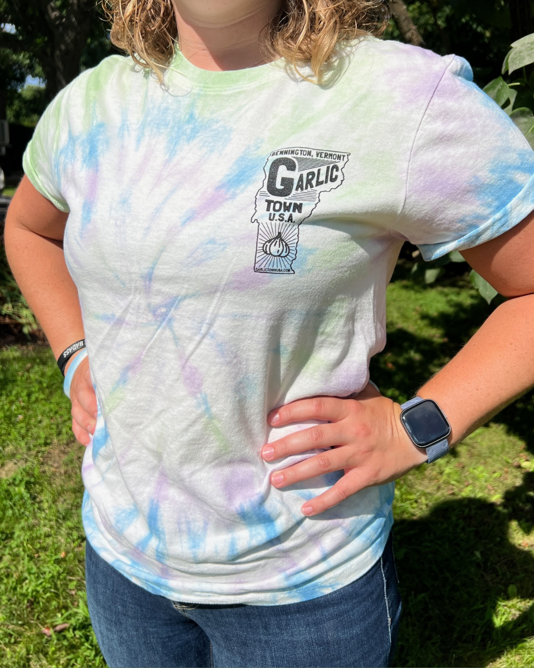 Close-up of a person wearing a tie-dye T-shirt with a graphic design and text about Bennington, Vermont and garlic. The person has a smartwatch on their left wrist and a black wristband on their right wrist, standing outdoors with greenery in the bac