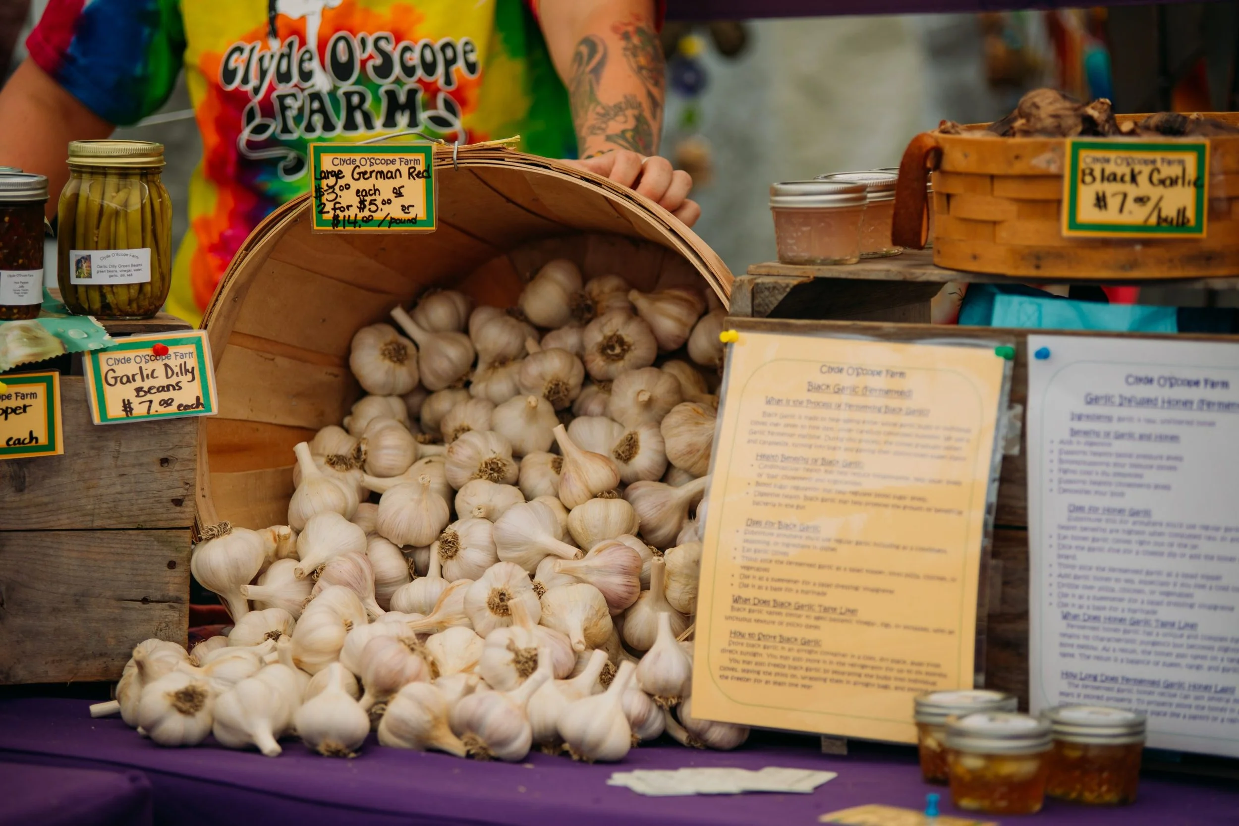 A display of fresh garlic bulbs at a farmer's market stall. The garlic is in a large woven basket with a sign indicating the price for German Red garlic. There are jars of pickled garlic or related products nearby, along with handwritten signs and in