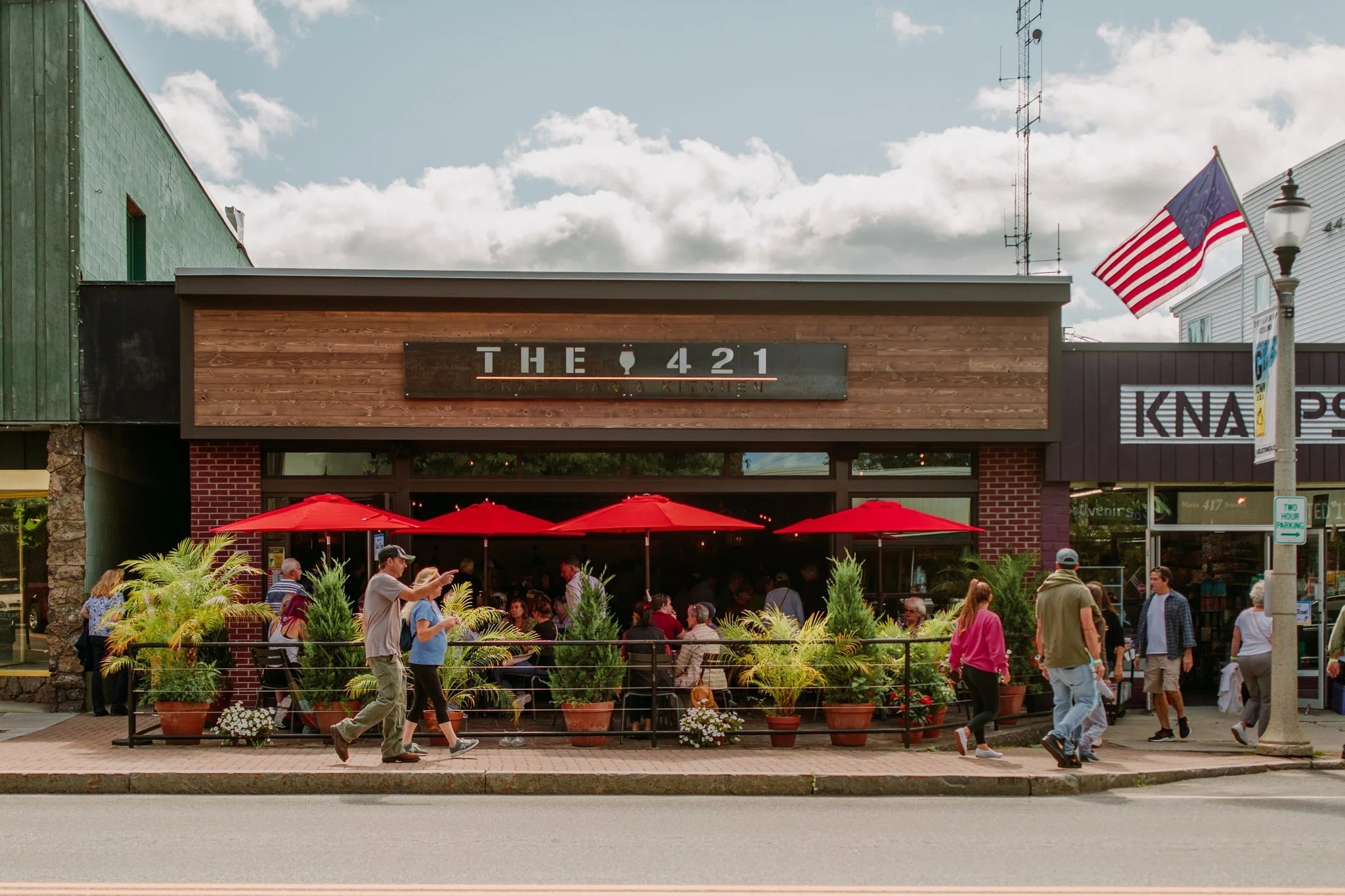 People dining and walking outside of a restaurant called THE 421 with red umbrellas, plants, and a glass facade on a busy street.