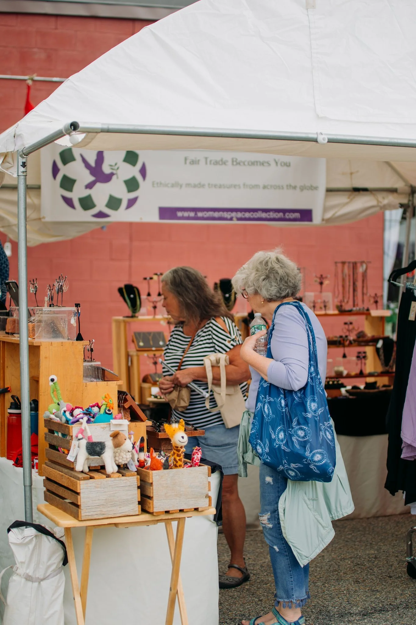 Two women browsing handmade jewelry and felt animal toys at an outdoor market stall, with a sign reading "Fair Trade Becomes You" and a pink brick wall in the background.