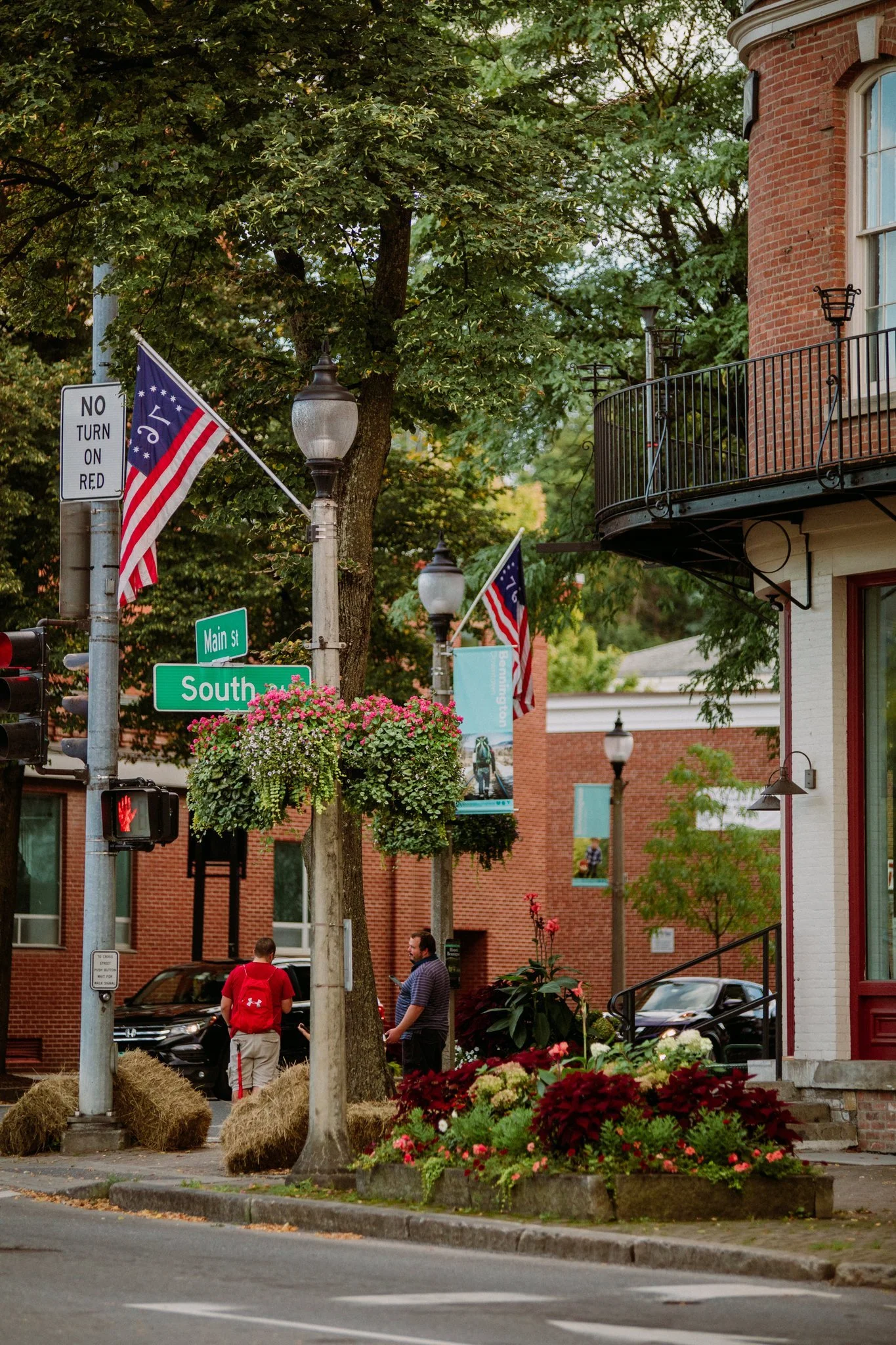 Street scene with American flags, street signs for Main Street and South, hanging flower baskets, a lamppost, two men conversing, parked cars, and a building with a balcony and decorative flowers.