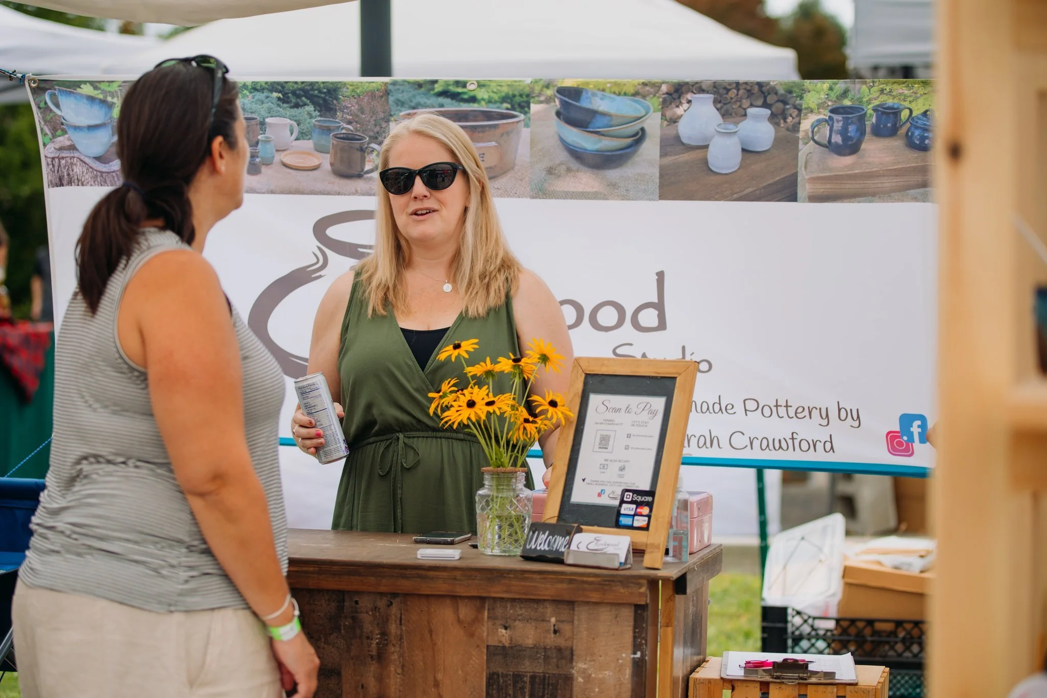 A woman with blonde hair, sunglasses, and a green dress is talking to a customer at a pottery booth. The booth has a sign that indicates handmade pottery by Sarah Crawford, with pictures of pottery items in the background and some flowers on the coun