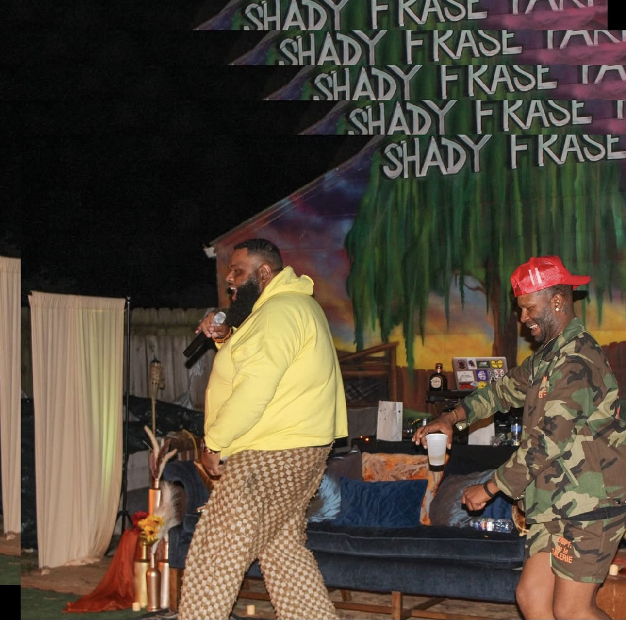 Two men are seen at an outdoor event, one wearing a yellow hoodie and patterned pants, singing with a microphone, the other in camouflage clothing and a red cap, holding a drink. The backdrop features a colorful mural with the words 'Shady F Rase' repeated multiple times.