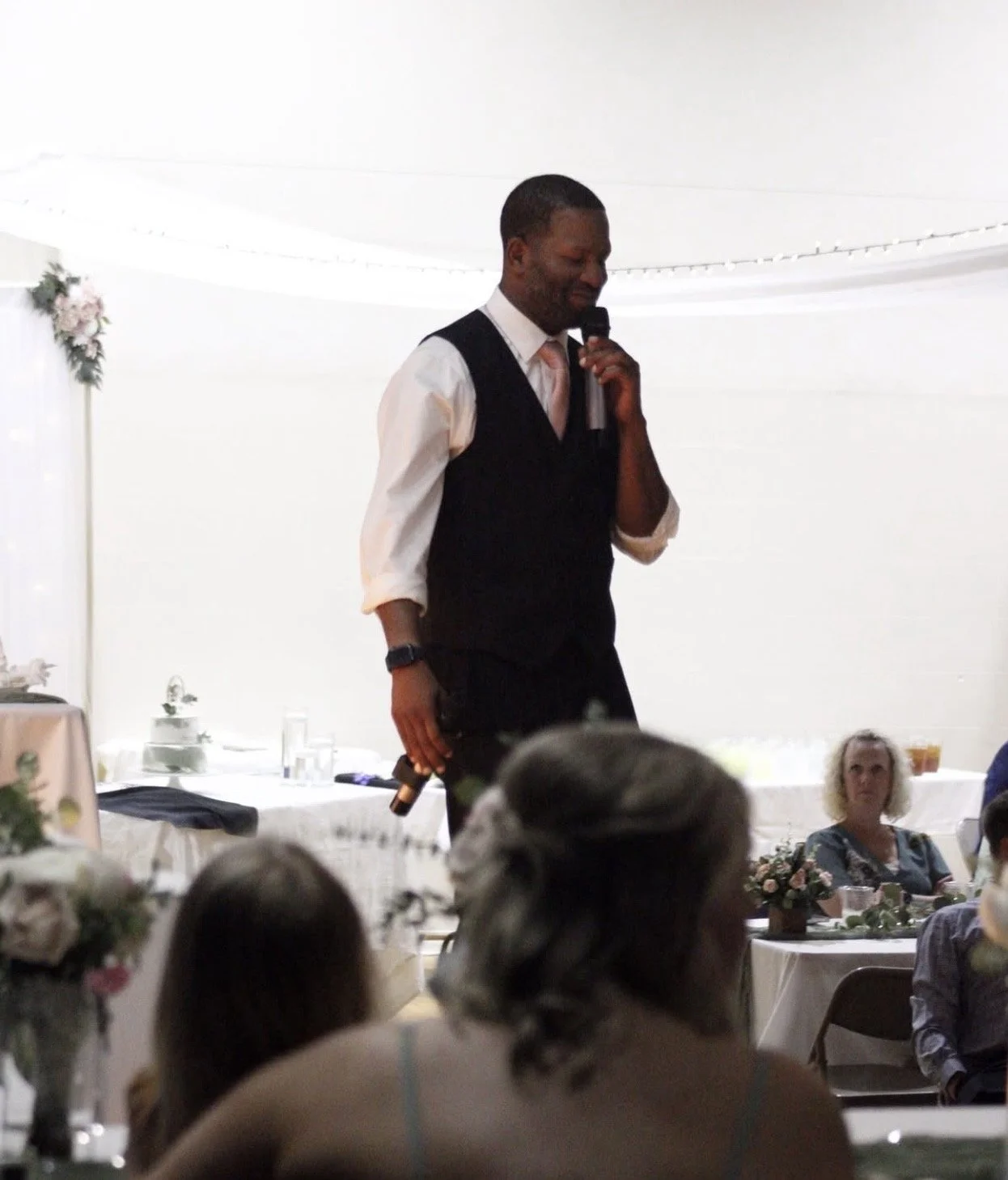 Man in a suit vest and white shirt holding a microphone, standing with his eyes closed, at a celebration or wedding reception with guests seated at decorated tables.