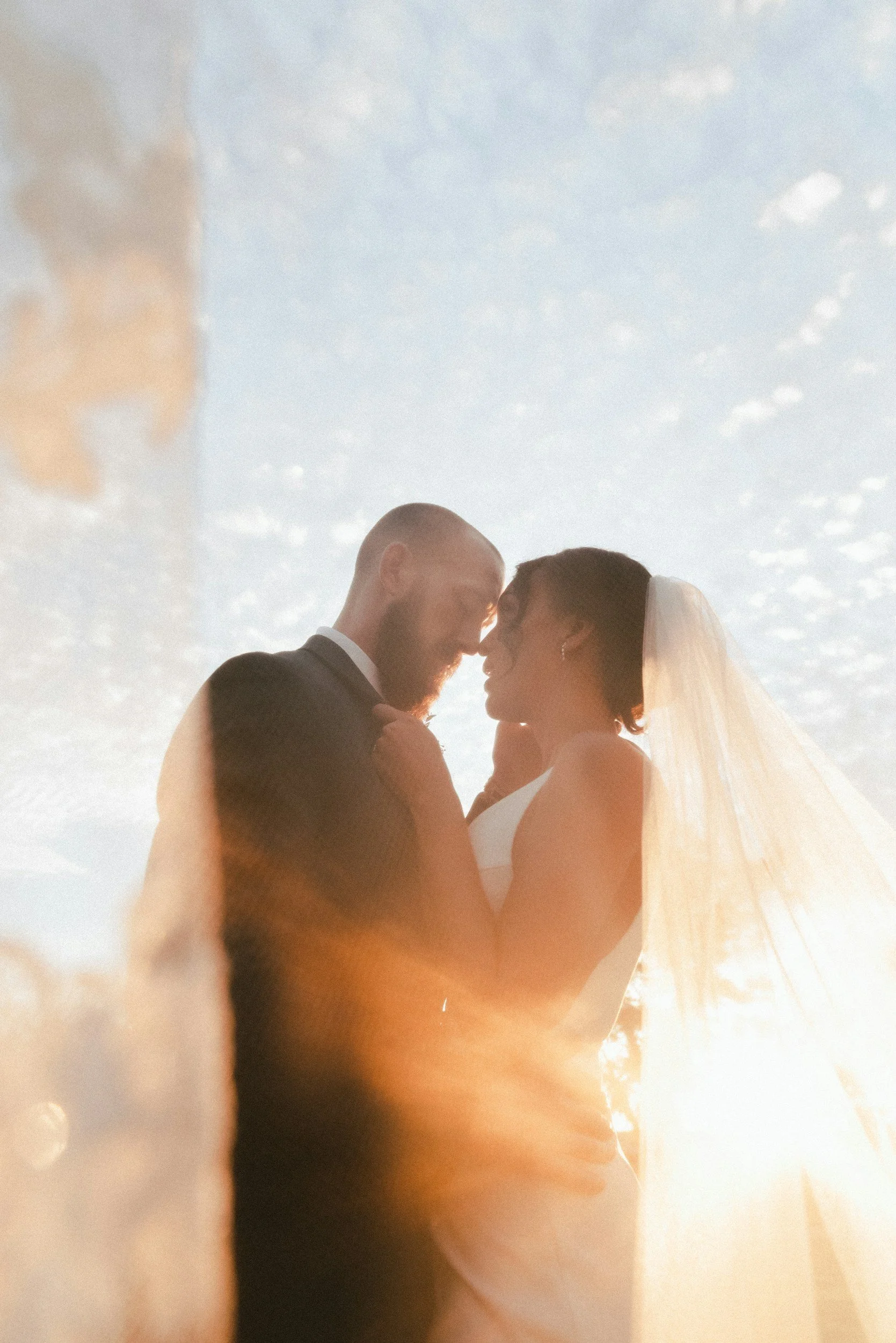 A bride and groom face each other outdoors during sunset, with their foreheads touching and eyes closed, illuminated by the warm sunlight and a partly cloudy sky in the background.
