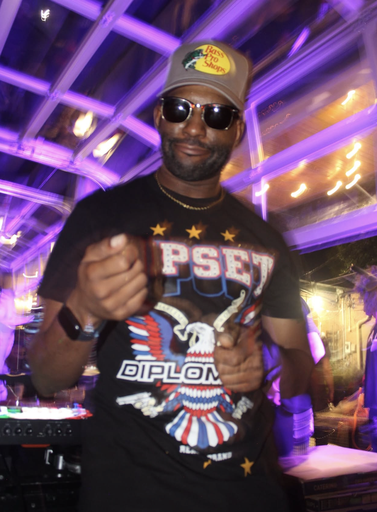 A man wearing sunglasses, a gray bass pro shops cap, a black t-shirt with patriotic eagle and star designs, and a watch, pointing at the camera at a lively event with purple and yellow lighting.