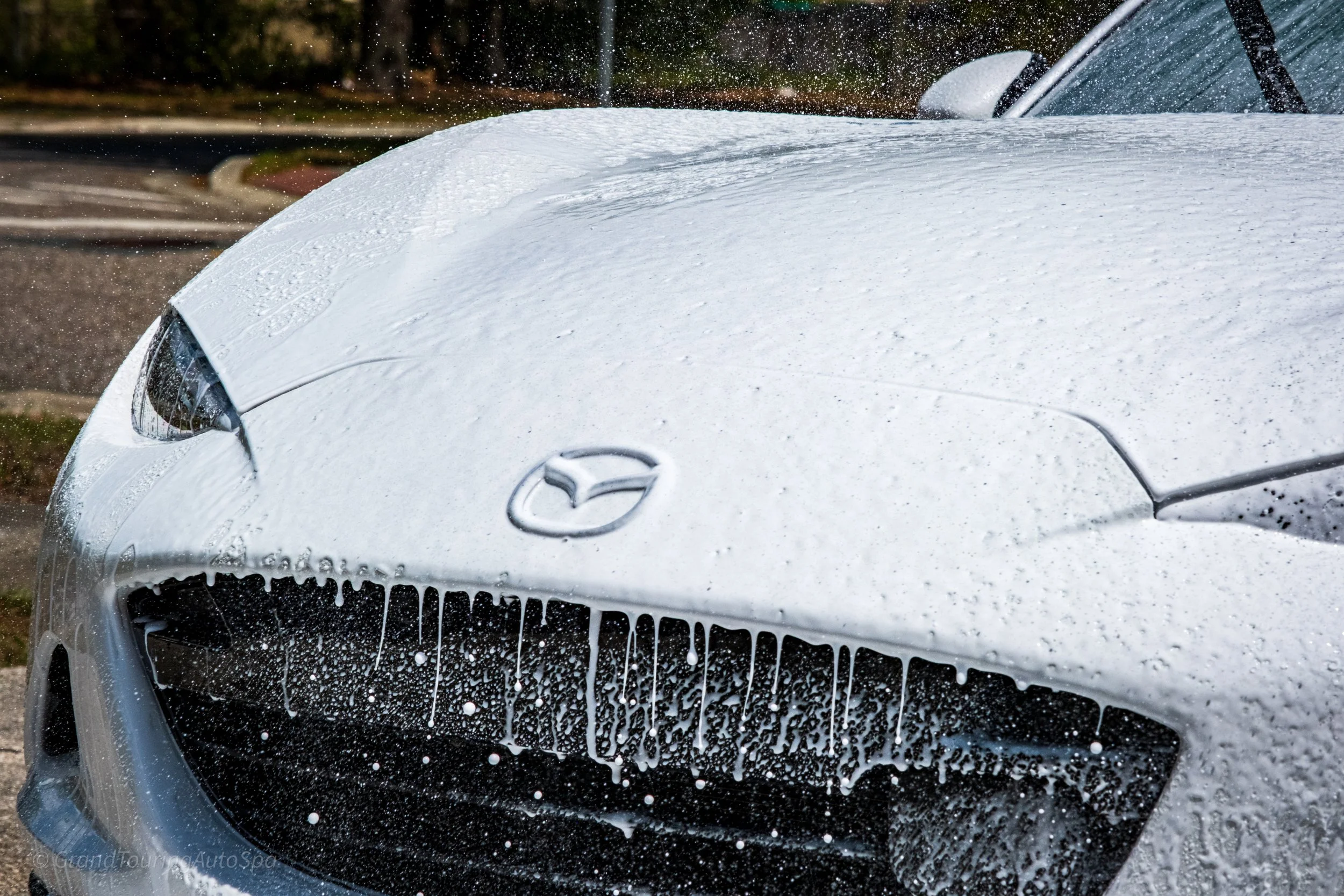 Close-up of a white Mazda car being sprayed with foam soap during a car wash.