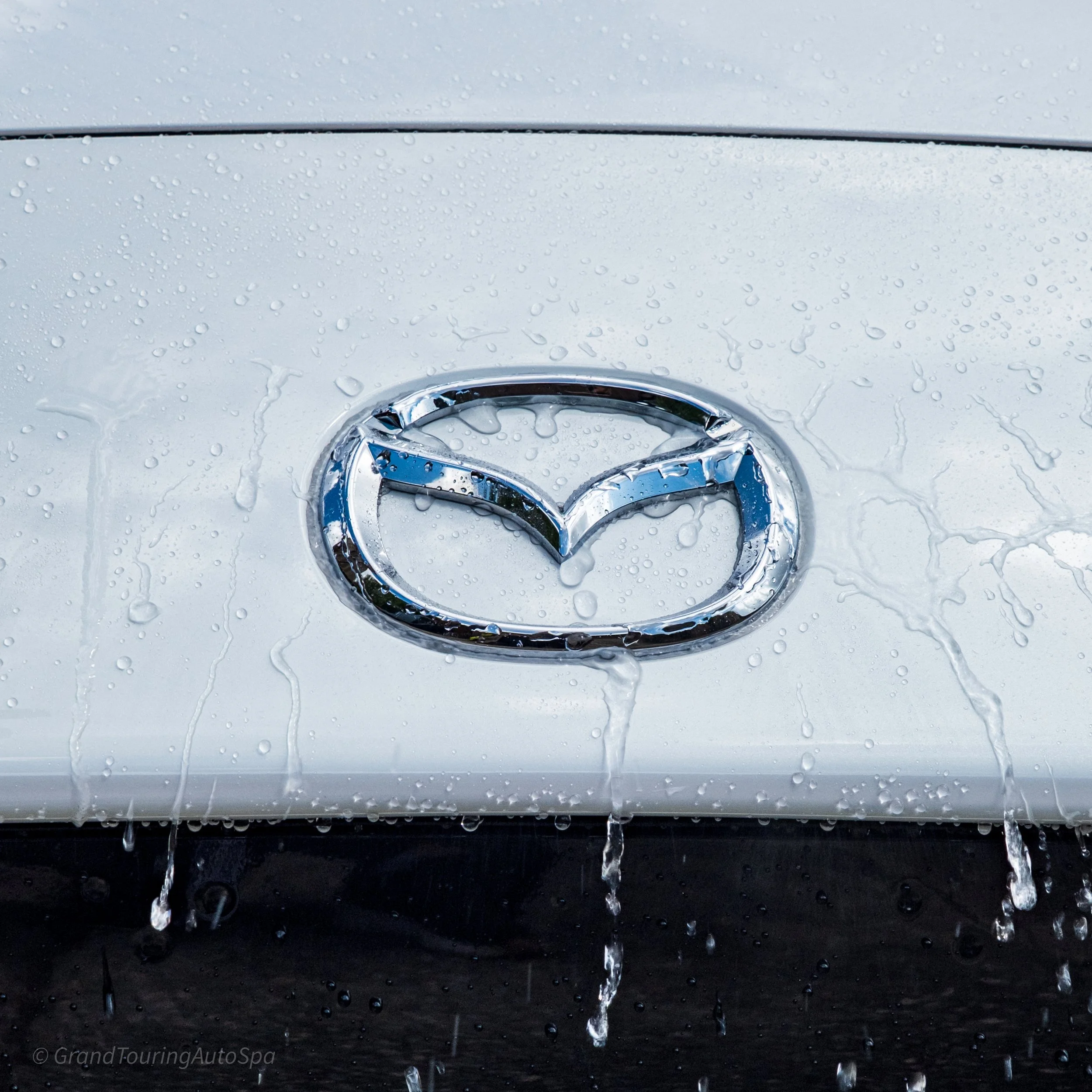 A Mazda emblem on a silver car, with water droplets on the surface and water dripping from the emblem.