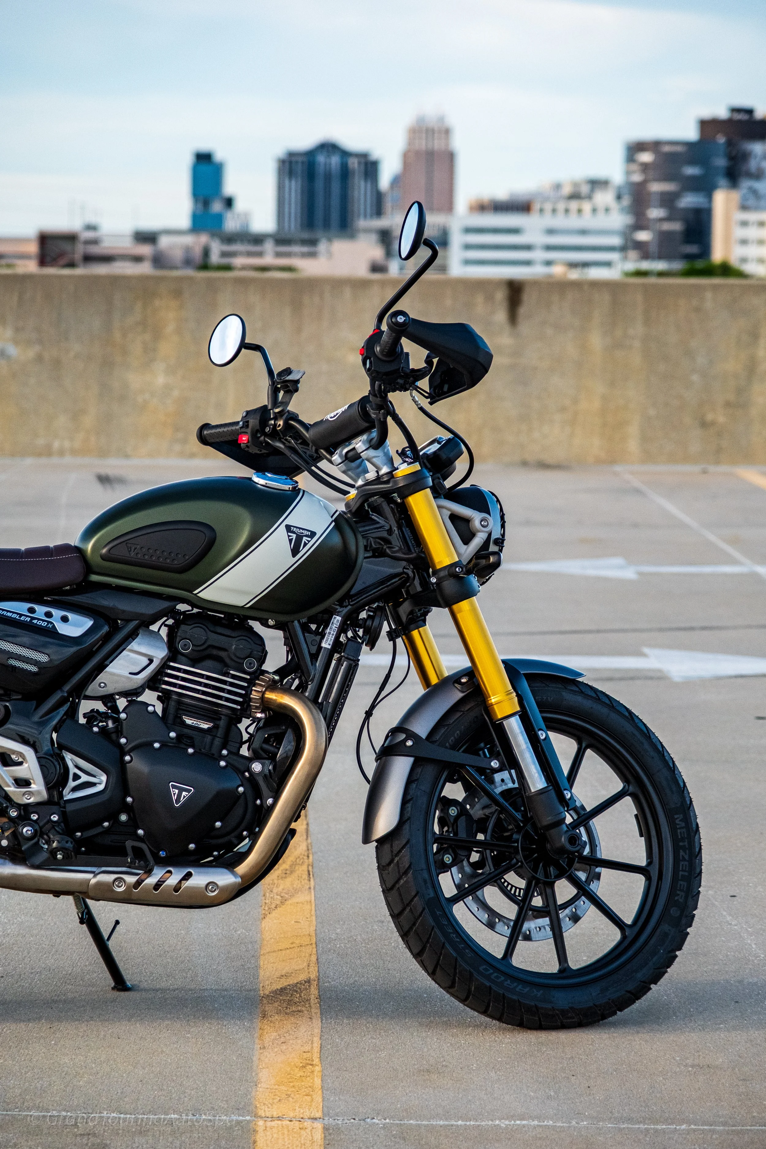 A Triumph motorcycle parked in an empty rooftop parking lot with a city skyline in the background.