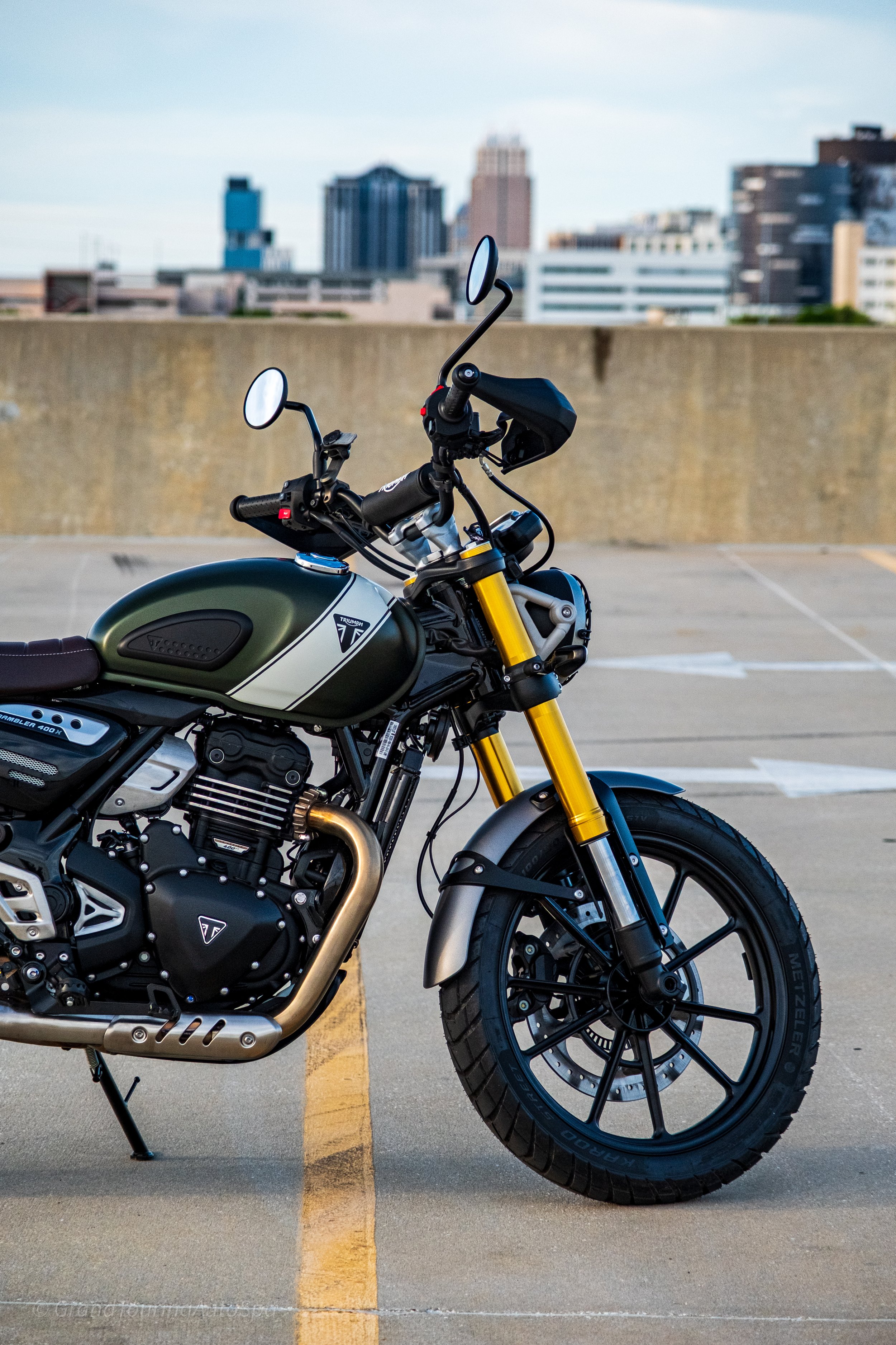 A green and black Triumph motorcycle parked on a rooftop parking lot with city buildings in the background.