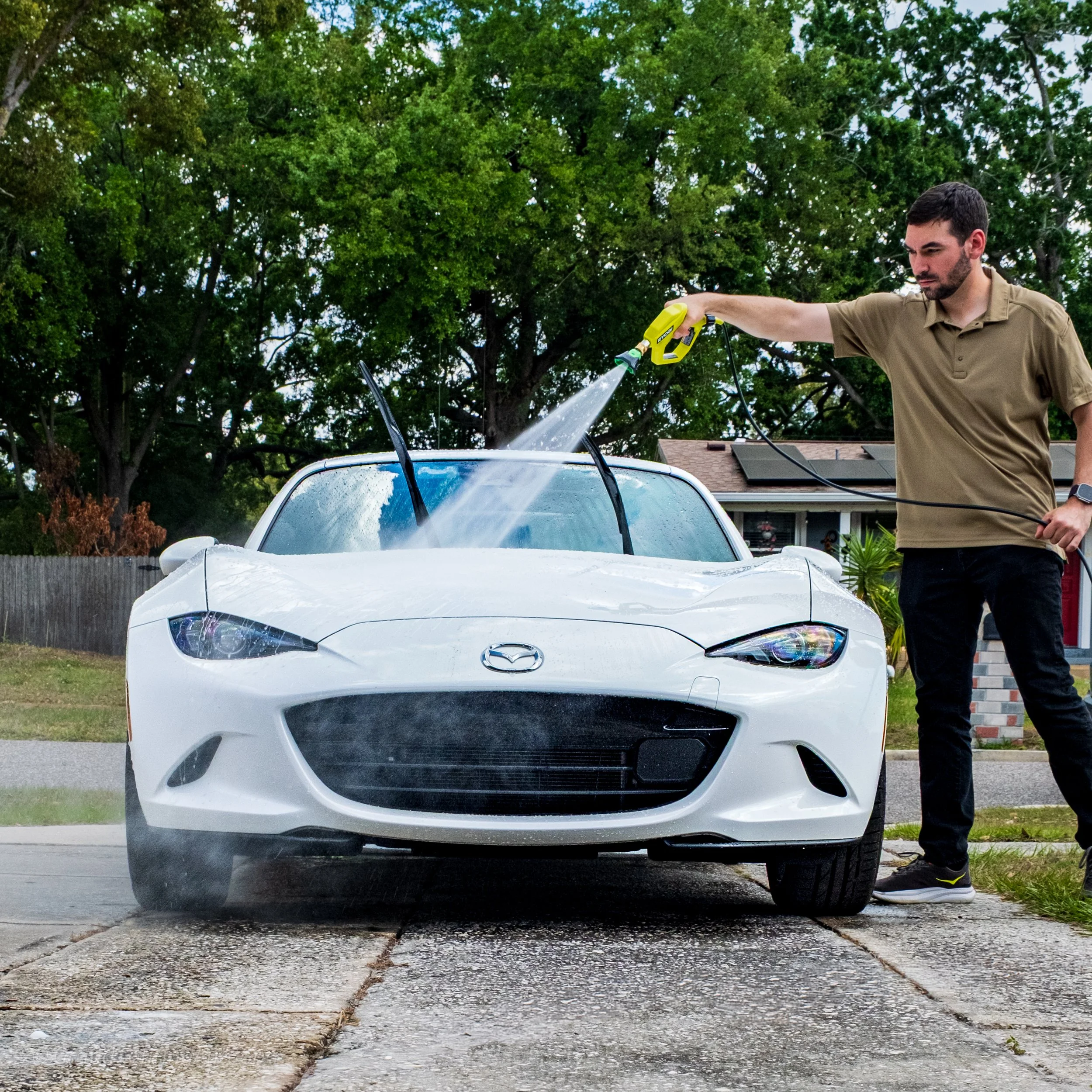 Man washing a white Mazda sports car with a garden hose on a driveway in front of a house with trees and a wooden fence.