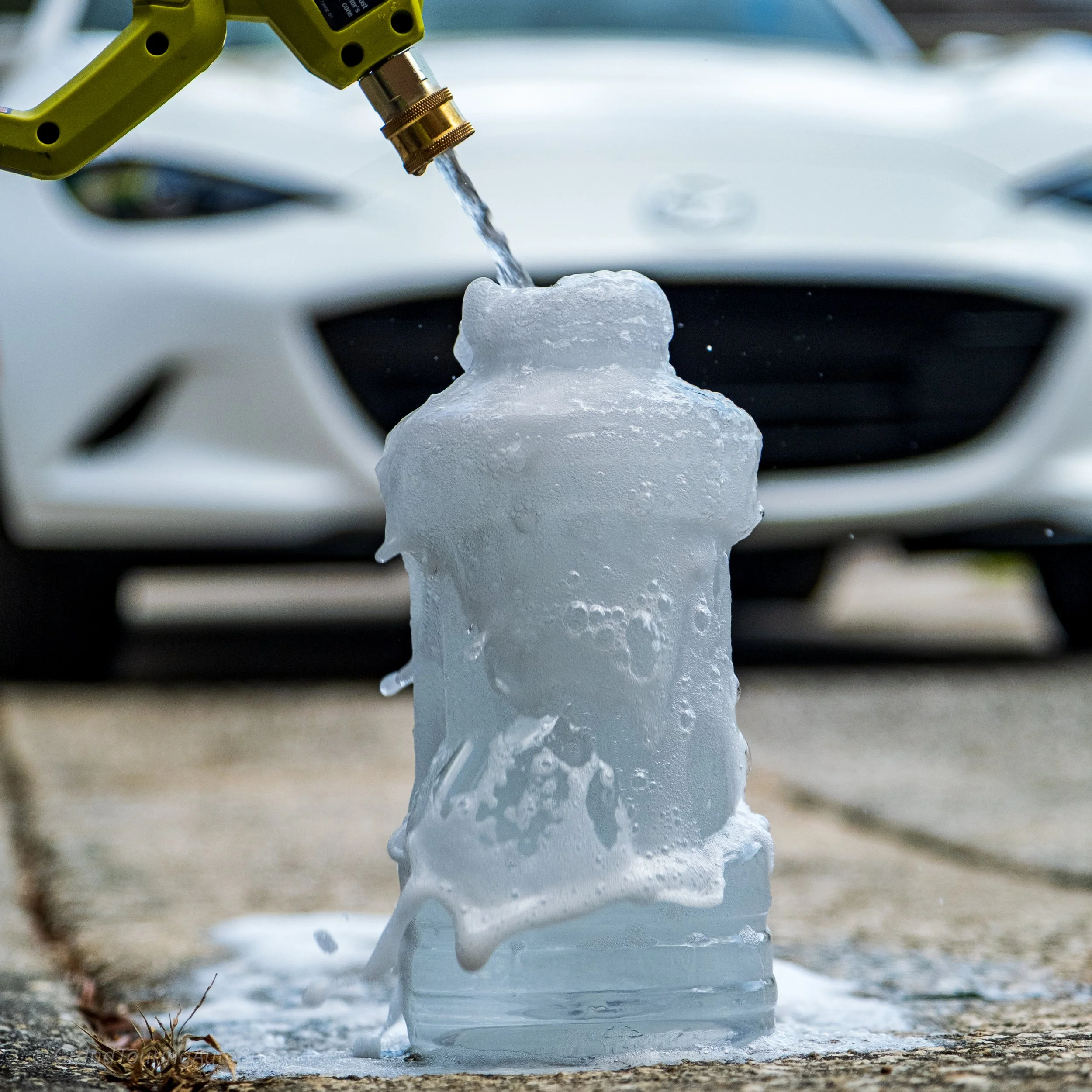 A large ice block is melting under a garden water hose with running water, with a white car in the background on a paved surface.