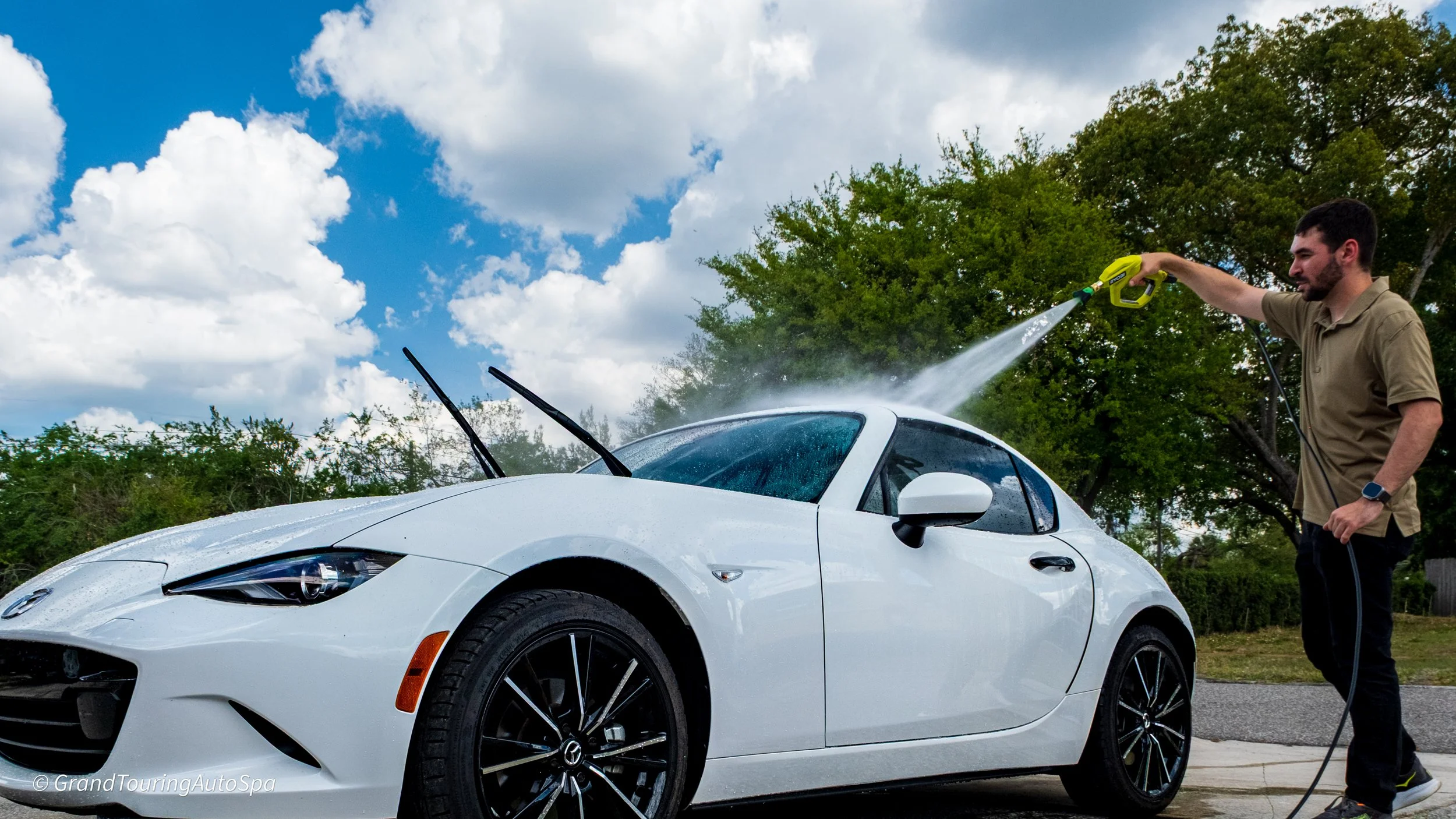 Man washing a white Mazda Miata convertible car with a pressure washer on a driveway with trees and cloudy sky in the background.