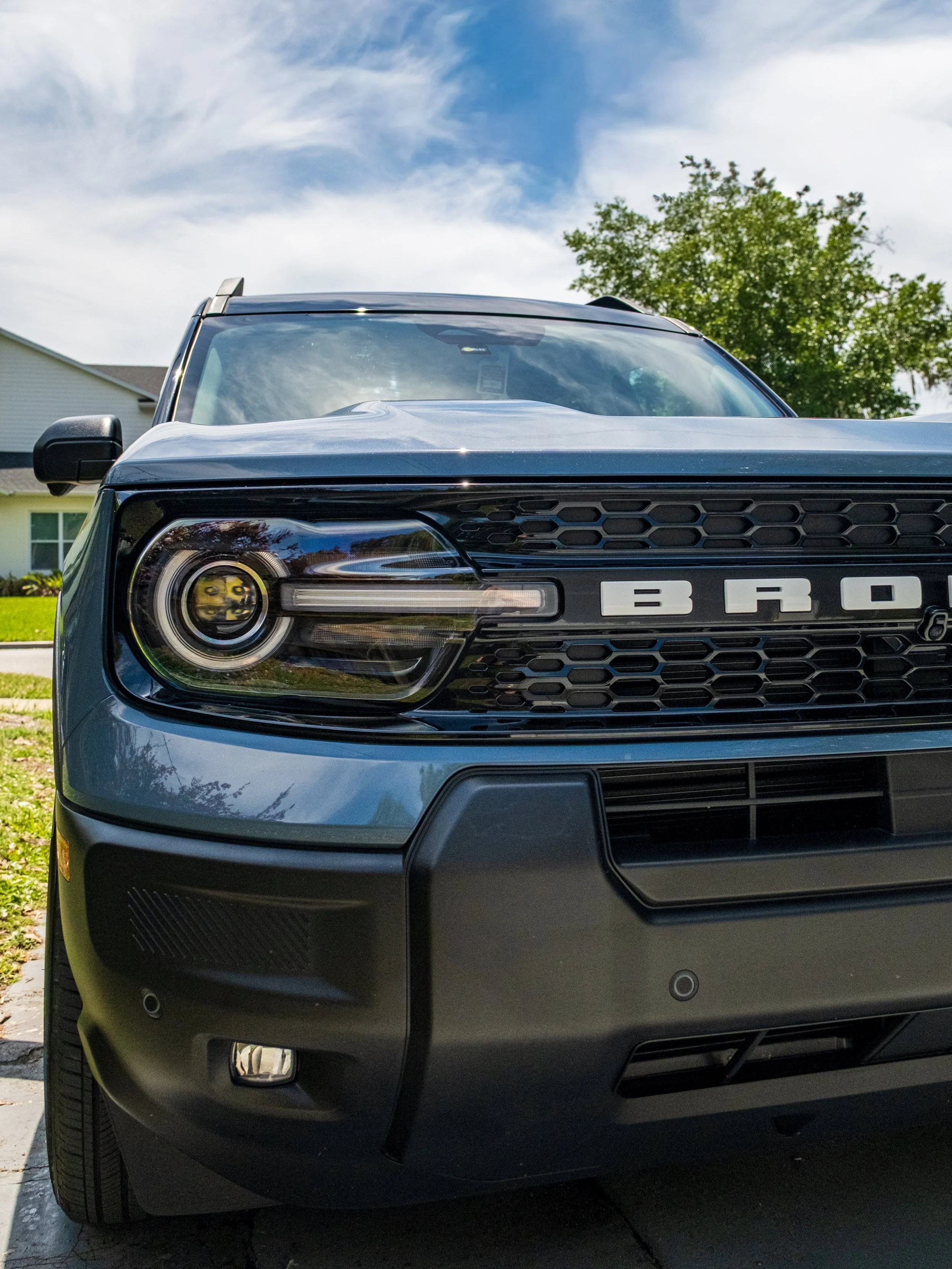 Front view of a black Ford Bronco parked outdoors on a sunny day with houses, trees, and a blue sky with clouds in the background.