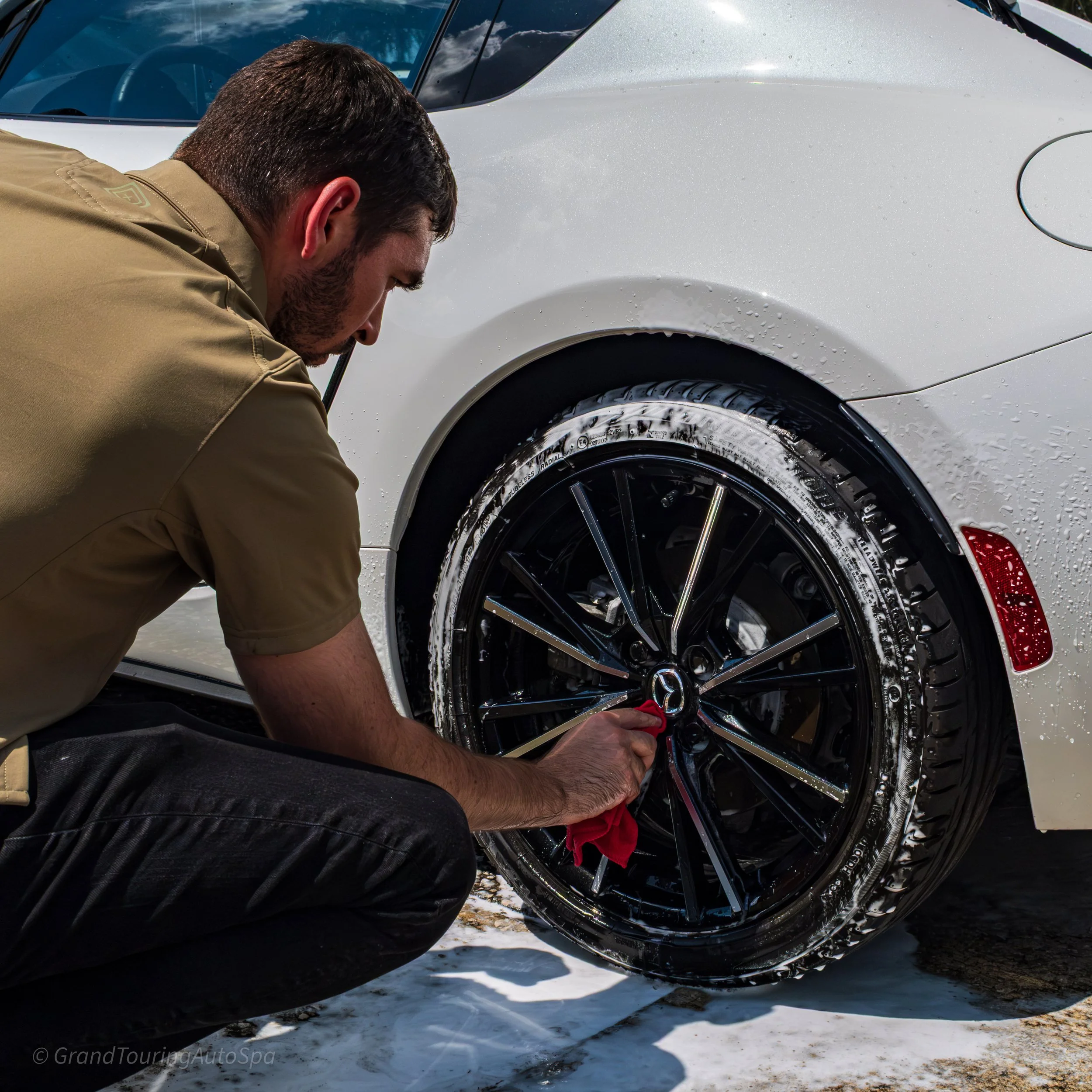 A man cleaning the black wheel of a white car during a car wash, with soap and water on the tire and wheel.