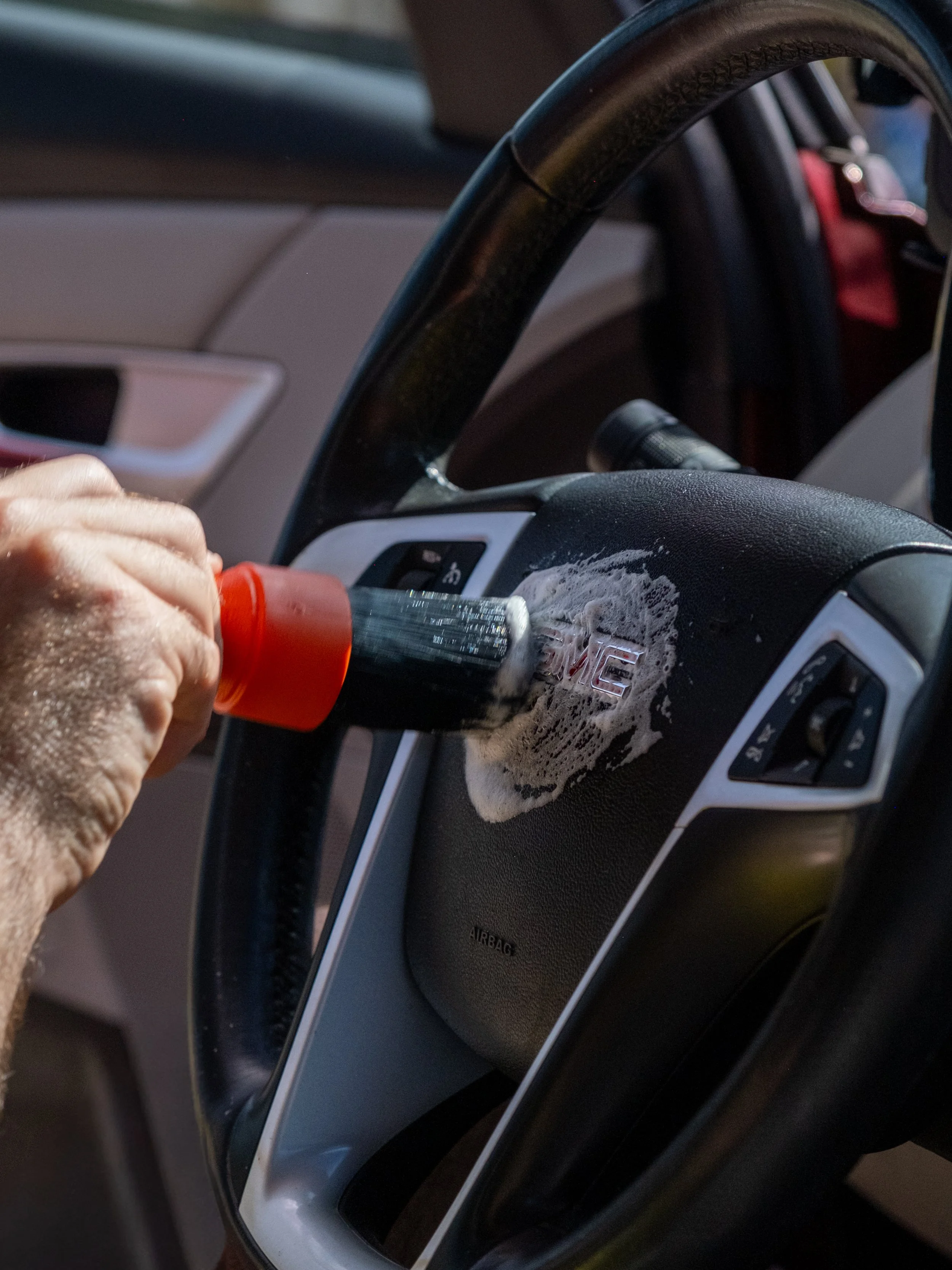 A hand holding a scrub brush cleaning the steering wheel of a car with soap suds.
