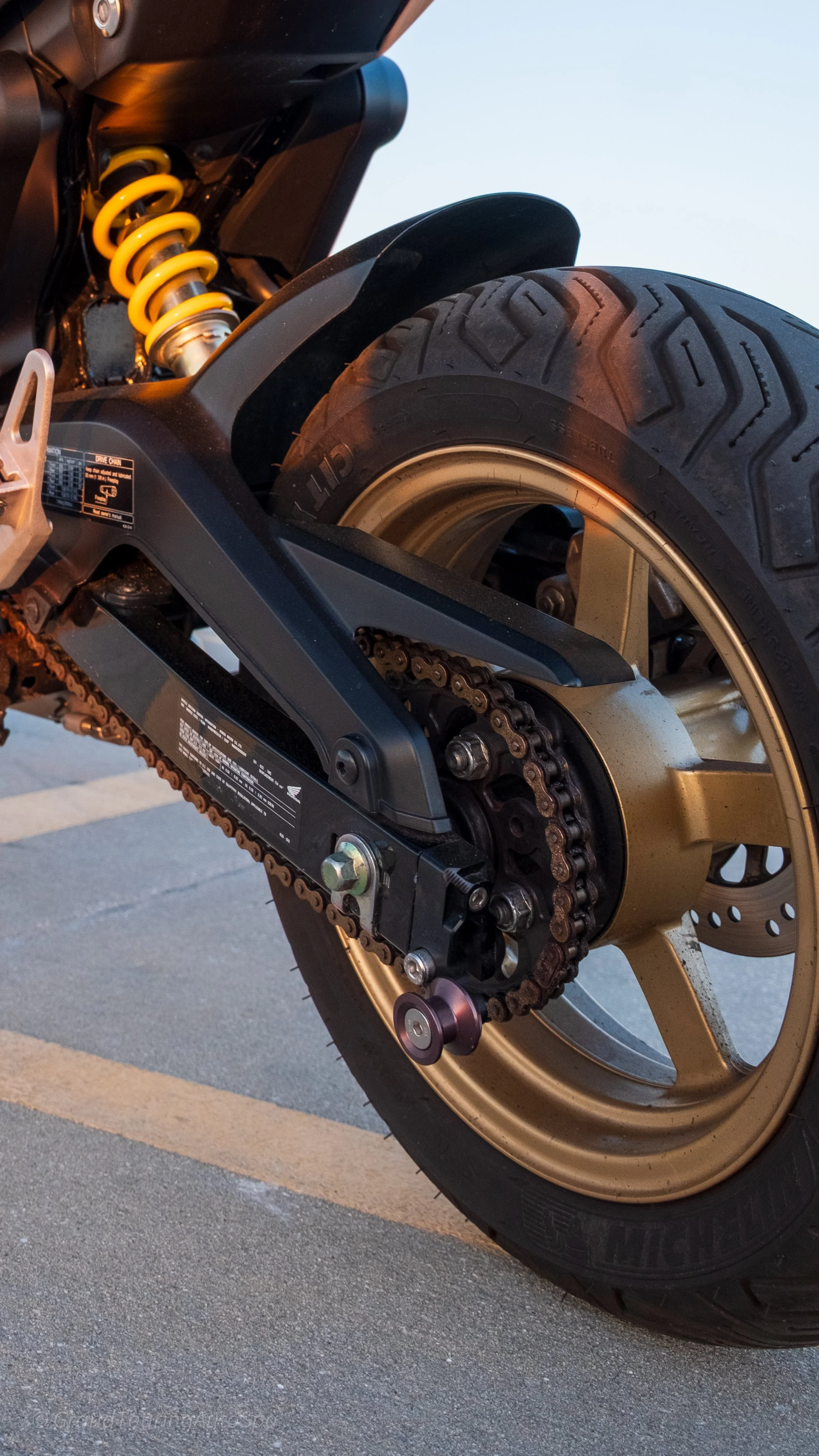 Close-up of a motorcycle rear wheel, chain, and suspension system parked on a paved surface.