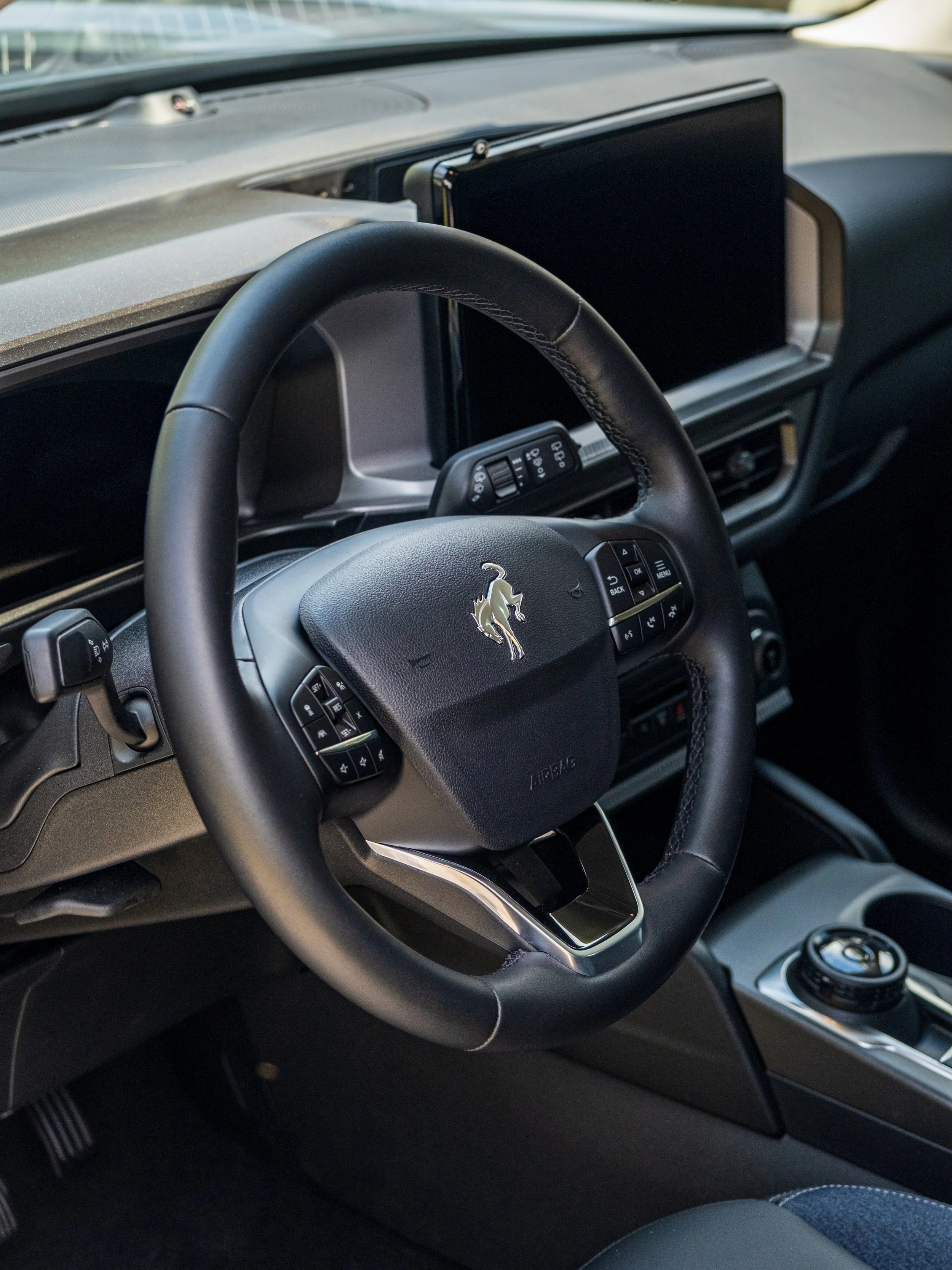 Inside view of a modern car dashboard featuring a steering wheel with a logo, a digital display screen, and control buttons, with the gear shift and other interior elements visible.