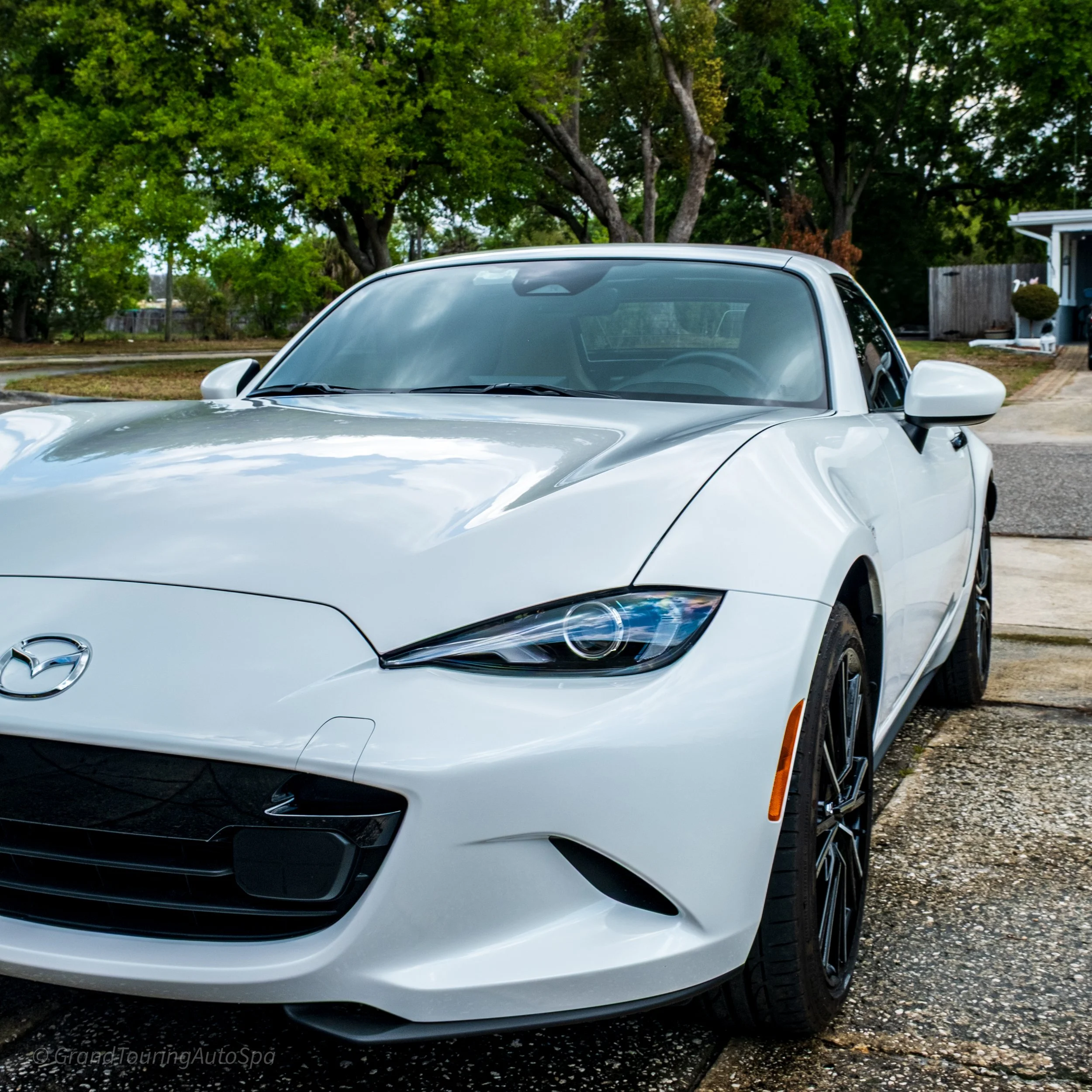White Mazda convertible car parked on a driveway with green trees and a house in the background.